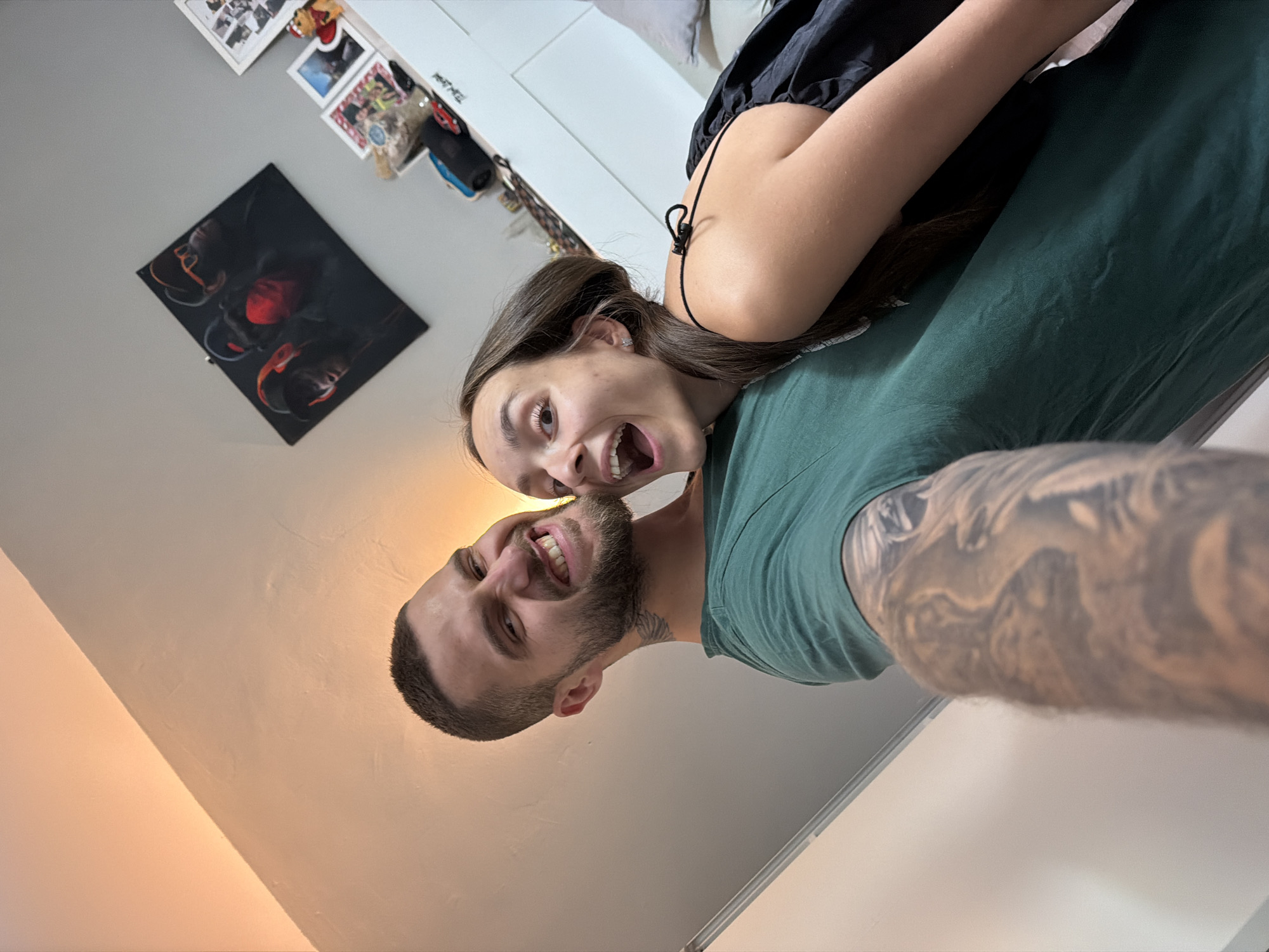 In the city of Sofia, Bulgaria, a man with short hair and a beard, wearing a green t-shirt and adorned with tattoos on his arm and neck, is taking a selfie. He is looking at the camera with a wide, playful grin, his teeth showing. Beside him, a woman with long, dark hair is leaning into him, her mouth open in a surprised or excited expression. She is wearing a black dress with thin straps. They appear to be in an indoor setting, possibly a living room, with a wall and a piece of art featuring three figures in the background. The lighting suggests it might be evening.