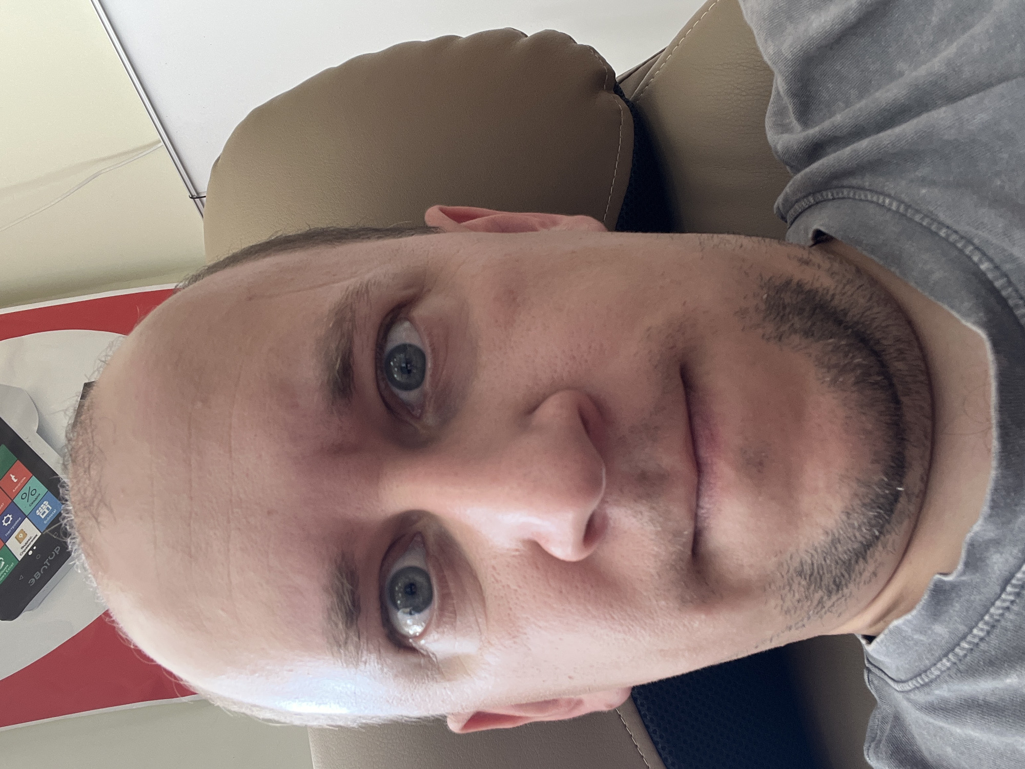 This is a close-up, self-portrait photograph of a Caucasian male in his late 20s to early 30s. He has fair skin, a receding hairline with short, light brown hair, and visible stubble on his chin and jawline. His eyes are wide open and a striking blue-gray color, and his gaze is directed at the camera. He is wearing a gray, crew-neck t-shirt.
The man appears to be seated in a chair with a brown, cushioned headrest. In the background, to the left and out of focus, is a red and white object with some text visible, possibly a logo or advertisement. To the right of the man, the background is a plain, light-colored wall, with a metallic rod or strip running horizontally across it. The lighting in the image suggests an indoor setting.
There is no information within the image to indicate the specific location in Omsk, Russia.