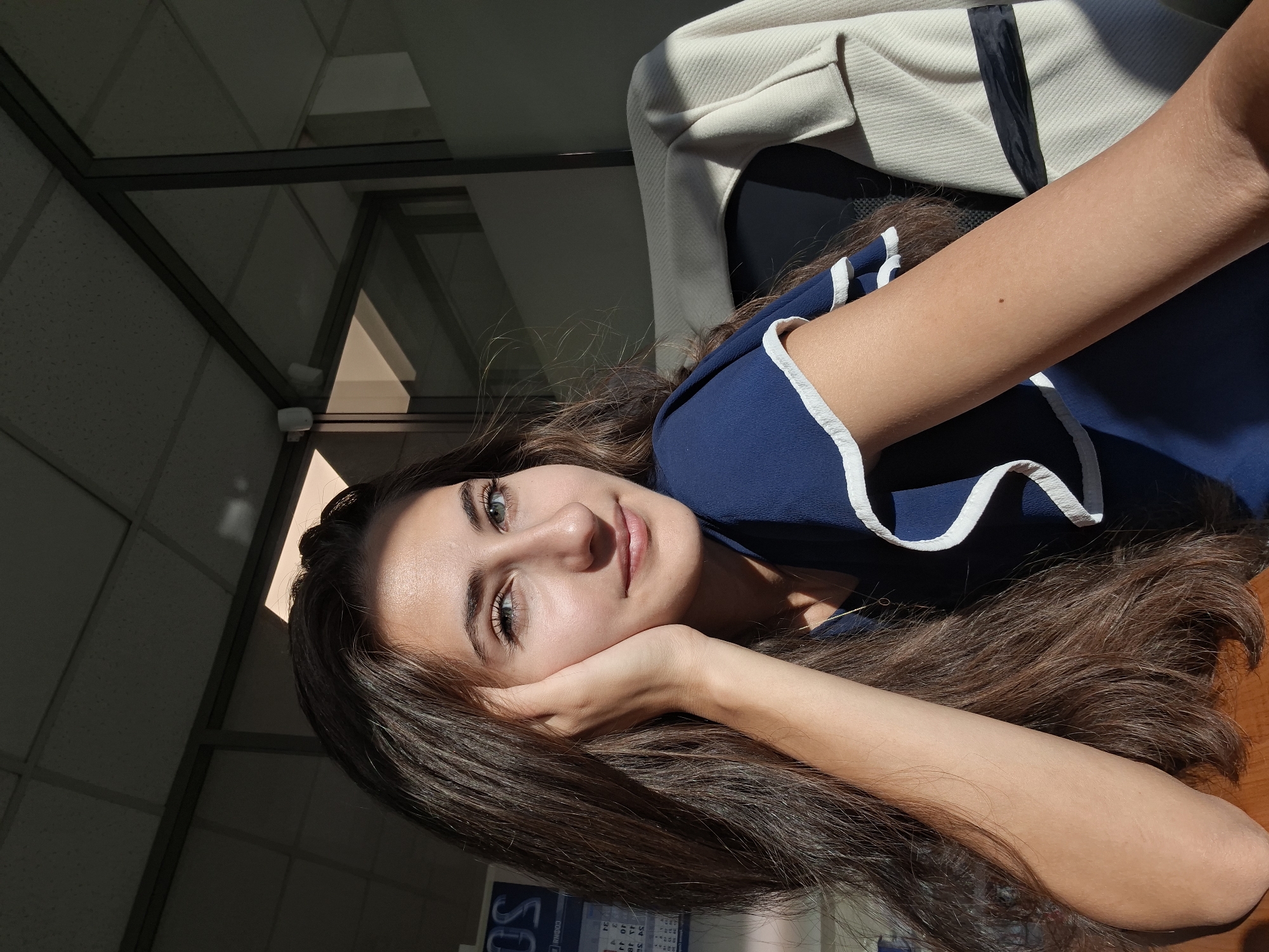 A medium shot captures a young woman from the chest up, seated indoors, with her left hand propped against her cheek. She has long, dark brown hair that cascades over her left shoulder, and her right arm rests on a wooden desk. She wears a dark blue top with white ruffled trim on the sleeves. Her eyes are a striking blue-green, and she is looking towards the camera with a gentle smile. The background suggests an office environment, with frosted glass partitions, a white ceiling with fluorescent lights, and a light-colored jacket draped over the back of her chair. Sunlight streams in from the left, casting distinct shadows on the ceiling and highlighting the woman's face and hair. A calendar is visible on the wall behind her to the left.