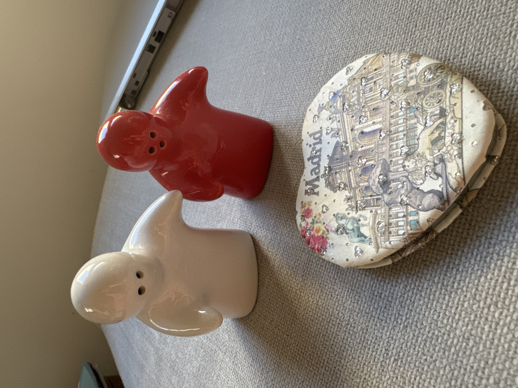 The image shows two small ceramic figures, one white and one red, resembling abstract people with simple facial features. In the foreground, a heart-shaped trinket box is decorated with a scene depicting buildings, animals, and flowers, with the word "Madrid" clearly visible on its surface. The objects are placed on a textured, neutral-colored surface, likely a couch or fabric. The background is out of focus, showing a wall and a thin, silver-colored object. There is no indication of the city of Murcia, Spain, in the image.