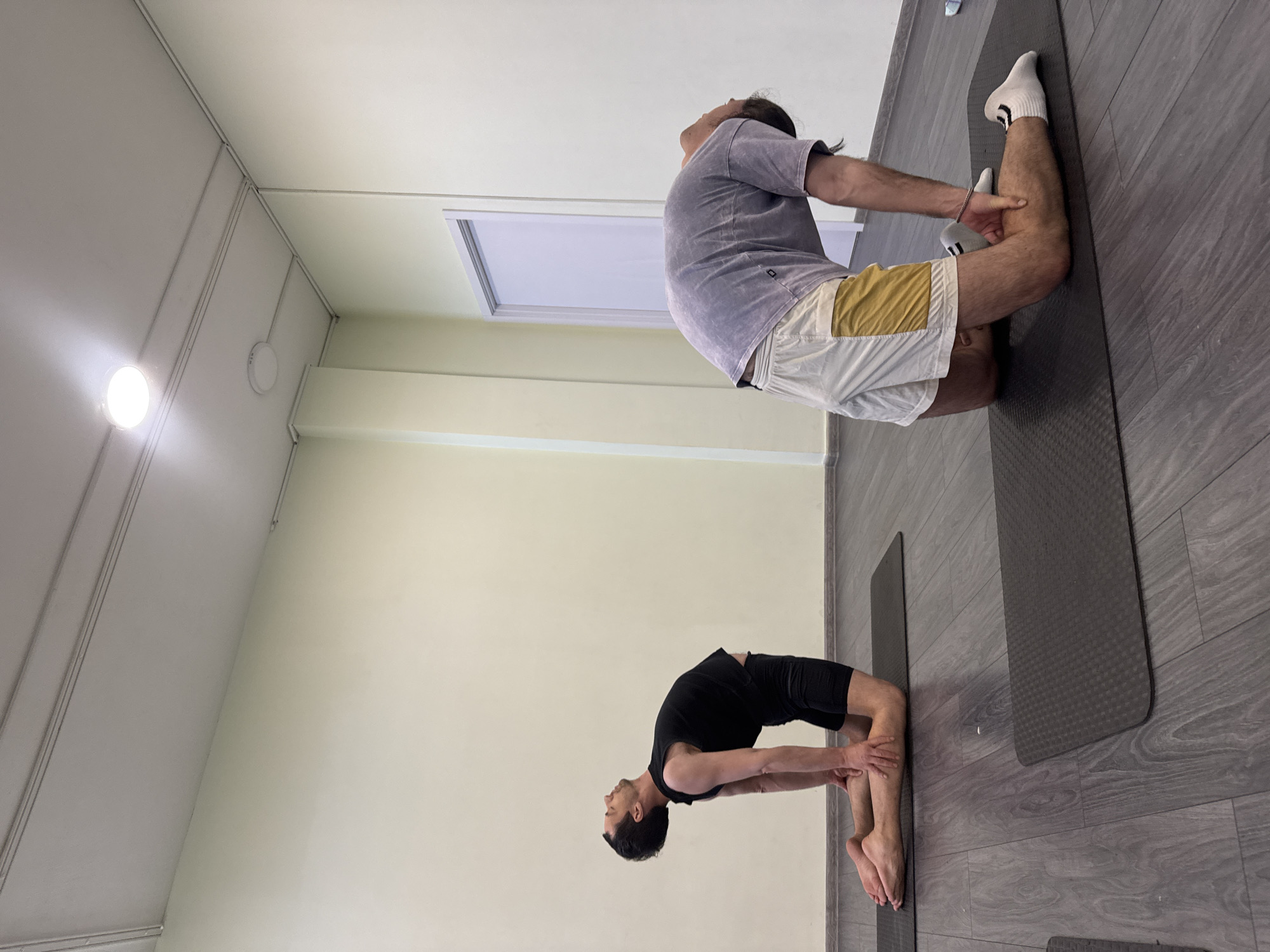 In an indoor setting, two individuals are engaged in a yoga pose known as Ustrasana or Camel Pose. The person on the left wears a black t-shirt and shorts, positioned on a yoga mat against a light green wall. The individual on the right is dressed in a purple t-shirt, white shorts with a yellow accent, and white socks, also on a yoga mat. The floor is a dark gray wood-look laminate. The room is illuminated by an overhead light, with a white door visible in the background. The location is Unknown, Unknown.