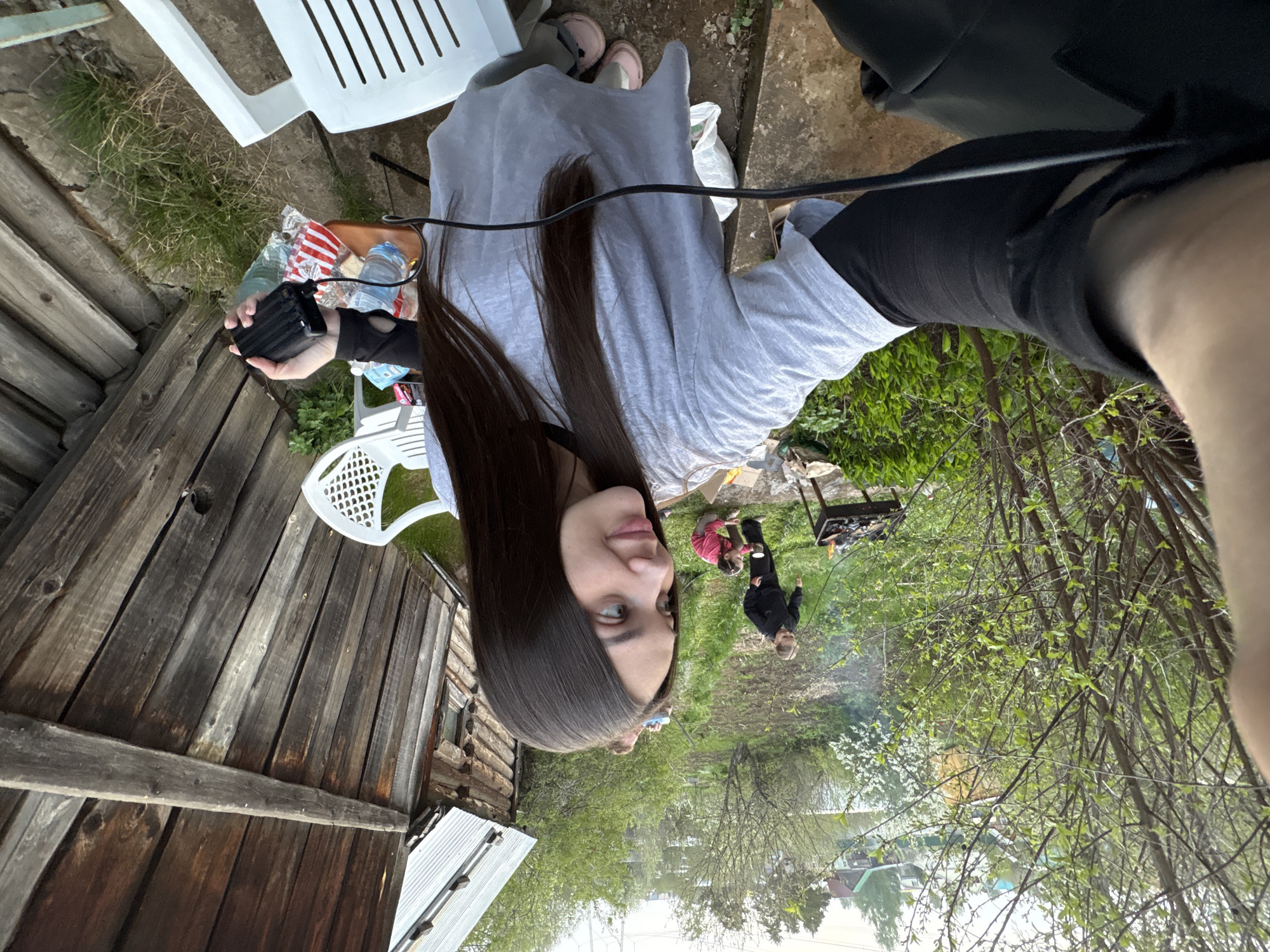 A woman with long, dark hair and light-colored eyes poses for a selfie. She is wearing a light-colored t-shirt and black pants, and holding a black object in her left hand. The background reveals an outdoor scene, seemingly in a backyard or garden setting. A wooden structure, possibly a shed or barn, is visible on the left side of the frame. In the distance, two people appear to be attending to a barbecue grill. Trees with green foliage are scattered throughout the background. The image location is in the city of Chaikovsky, Russia.