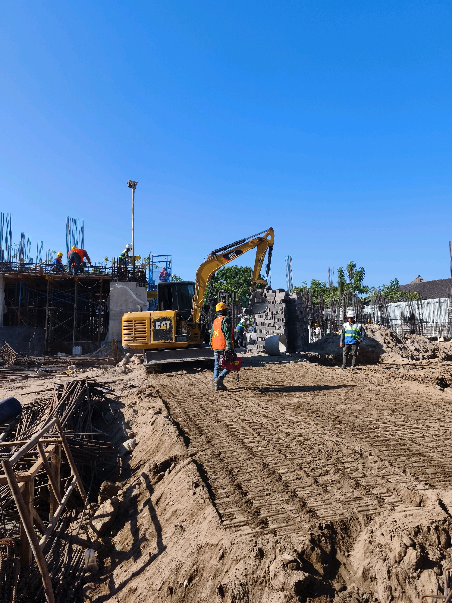 This is a photograph of a construction site in Denpasar, Indonesia, bathed in bright sunlight under a clear blue sky. The scene depicts ongoing construction with several workers present. A yellow Caterpillar excavator is a prominent feature, positioned in the mid-ground. In the foreground, a pile of rebar and other construction materials is visible. Several workers, some wearing hard hats and high-visibility vests, are scattered across the site. One worker, wearing a blue shirt and an orange safety vest, is walking towards the right of the frame, carrying a red container. Another worker in a white hard hat and blue vest stands in the mid-ground. In the background, the skeletal structure of a building under construction is evident, with scaffolding and rebar. Trees and a distant building with a dark roof suggest the surrounding urban environment of Denpasar. The ground is predominantly dirt and sand, with tire tracks from heavy machinery visible in some areas. The overall impression is one of active development and labor.