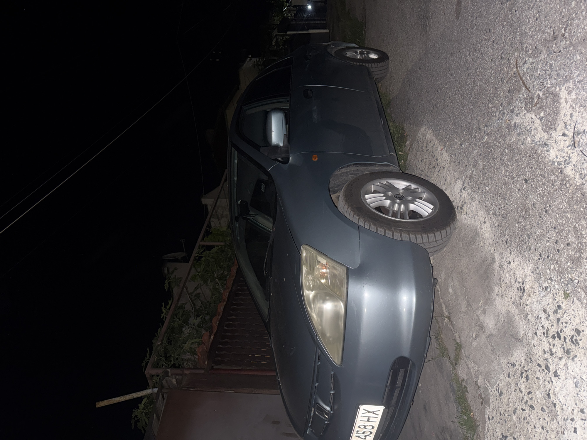 A dark gray car is parked on a concrete curb next to a patch of grass. The license plate visible reads "...458 HX." The street appears to be paved with asphalt. The image was likely taken at night. The city is Elhovo, Bulgaria.