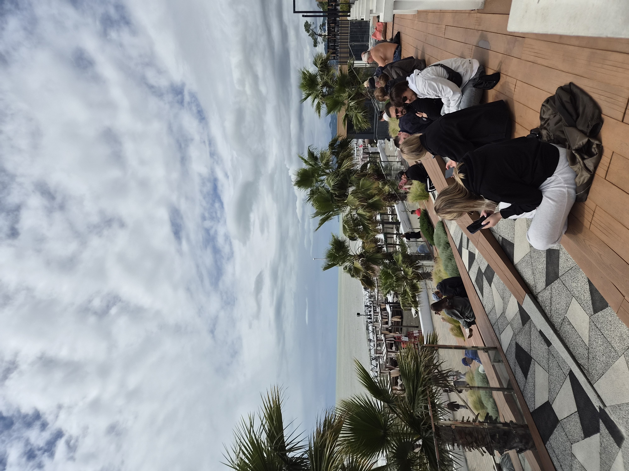 The image shows a gathering of people on a wooden deck, likely a promenade or rooftop terrace, overlooking a beach and the sea. The location is likely in the city of Sochi, Russia, given the coastal setting and presence of palm trees, consistent with the region's subtropical climate.
A group of individuals are kneeling or sitting along the edge of the deck, some appearing to look out at the view, while others are engaged with mobile devices. They are dressed in casual clothing.
The beach below is lined with rows of lounge chairs and umbrellas. The water appears calm and the sky is overcast with partly cloudy conditions. Palm trees are visible, adding to the coastal atmosphere.