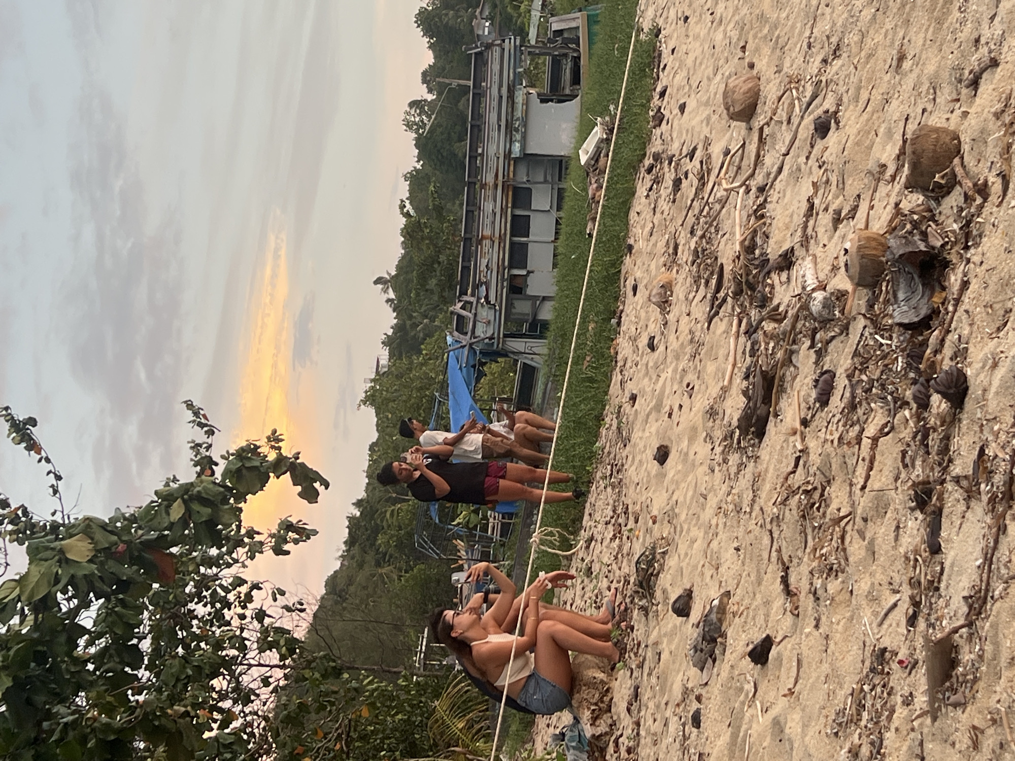The image depicts a beach scene in the Ko Pha-ngan District, Thailand, likely during the late afternoon or early evening, indicated by the soft, warm light of the sky and the muted tones of the landscape. The foreground is dominated by a sandy beach strewn with natural debris such as driftwood, dried leaves, and coconut husks. A white rope is visible stretched horizontally across the middle ground, separating the beach from the vegetated area behind.
On the left side of the image, a woman wearing a white halter top and denim shorts is seated on a large piece of driftwood. She is holding a mobile phone in her right hand and appears to be taking a selfie or a photograph. Her legs are crossed, and she is looking towards her phone.
Further back, a group of three people stands near a dilapidated boat. One person, wearing a black t-shirt and shorts, is holding a mobile phone up, possibly taking a picture or video. To their left, another person in a white t-shirt and shorts is also looking towards the phone or the direction it is pointed. A third individual, wearing a white t-shirt and dark shorts, stands slightly behind and to the right of the other two.
The background reveals a line of lush green vegetation, characteristic of a tropical environment, with trees and foliage. Beyond the vegetation, some structures are visible, suggesting a settlement or resort. A partially dismantled or abandoned boat, grey and weathered, is positioned behind the group of people and extends across a significant portion of the middle ground. The sky above is a soft gradient of pale blue and pinkish-orange hues, with wispy clouds, indicative of a sunset or sunrise. The overall impression is one of a casual, relaxed atmosphere on a tropical beach.