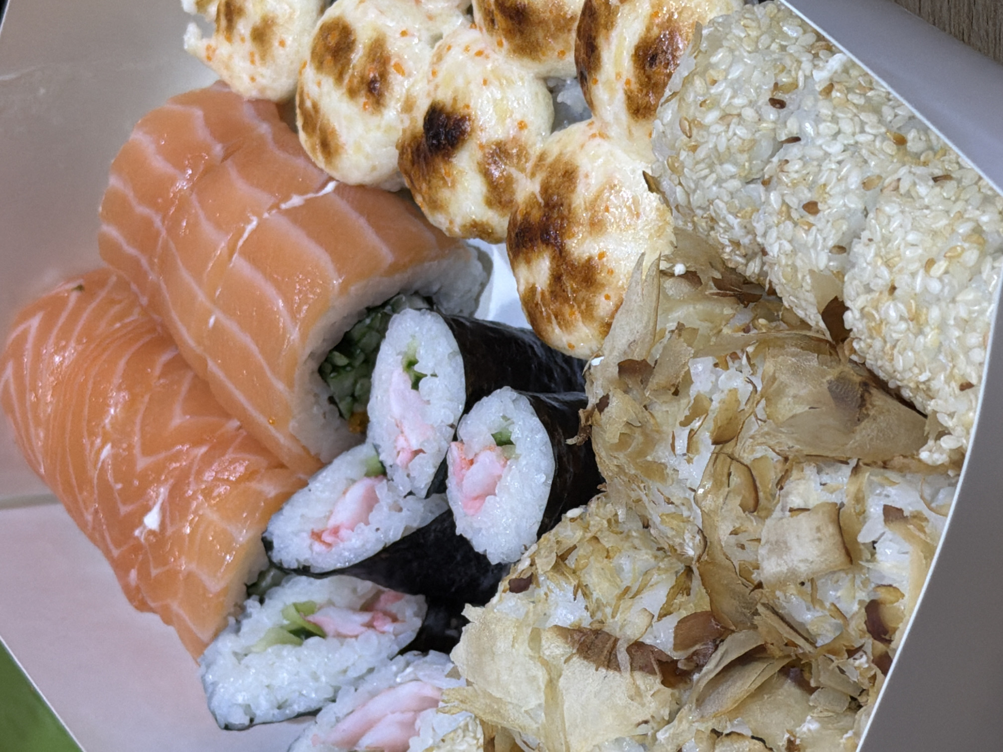 A top-down, close-up shot captures a white cardboard takeout box filled with a variety of sushi. The sushi includes several pieces of maki rolls topped with bright orange salmon, some of which are partially obscured by other elements. Next to these are round, bite-sized sushi pieces with a creamy, lightly browned topping, some speckled with orange roe or seasoning. In the foreground, there are two distinct sections of sushi. One is layered with what appears to be dried bonito flakes, giving it a textured, flaky appearance. The other section is covered in white sesame seeds, with some darker seeds mixed in. The lighting is natural and bright, highlighting the textures and colors of the food. The background is a soft, out-of-focus green. There are no people or any indication of the city of Khmelnytskyi, Ukraine.
