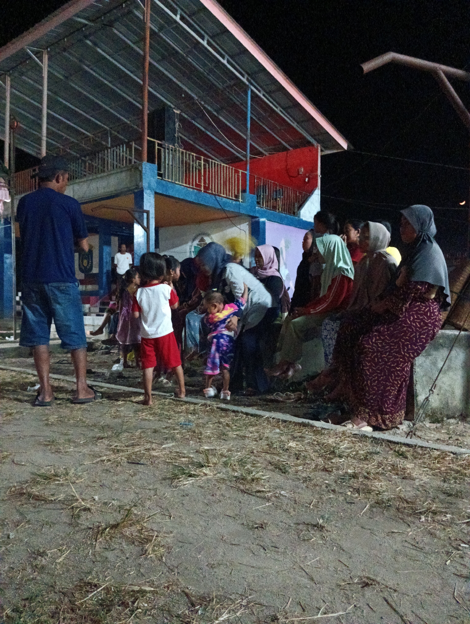 A group of people, including adults and children, are gathered outdoors at night, near a building with a covered roof. A man wearing a dark blue t-shirt and denim shorts stands on the left, facing away from the camera. Several women are seated on a low wall on the right side of the frame, some wearing hijabs. Children are visible in the center of the image, with one child in the foreground wearing a white t-shirt and red shorts. The ground is covered with dry grass and dirt. The scene appears to be in an open area, possibly a community space or event venue. The building behind the group has a multi-level structure with balconies and a sloped, metal roof. The lighting suggests it is nighttime, with artificial lights illuminating the area. The location is identified as Pematang Jaya, Indonesia.
