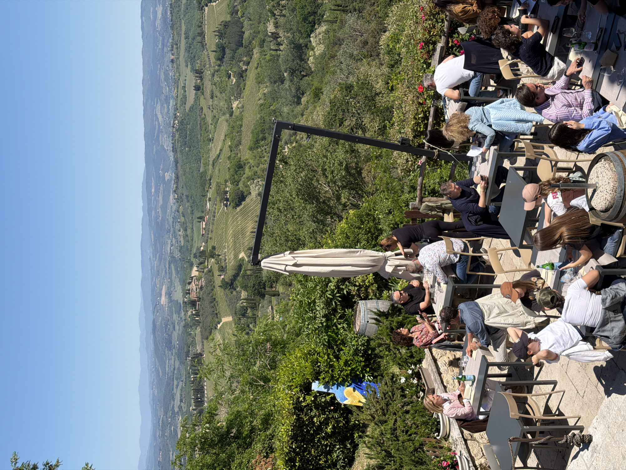 The image presents a vibrant outdoor dining or social gathering scene on a patio overlooking the panoramic Tuscan countryside, identified as being near San Gimignano, Italy. Numerous adults are seated at rectangular tables or standing, engaged in conversation, dining, and drinking. Many are dressed in casual to smart-casual attire. Tables are equipped with glasses and bottles, some clearly identifiable as green San Pellegrino water bottles.
The patio itself is paved and features a large, light-colored patio umbrella offering shade, as well as a prominent black metal structure, possibly part of an awning or architectural element. Lush green bushes, flowering plants with red and white blossoms, and a distinctive large blue and yellow ceramic vase or sculpture adorn the edges of the dining area. A wooden barrel filled with small stones or gravel is also visible on the patio.
Beyond the immediate patio, a vast and picturesque landscape unfolds under a clear, bright blue sky. Rolling green hills covered in vineyards, dense forests, and scattered traditional Italian buildings forming distant villages characterize the scenic view. The bright sunlight and strong shadows suggest the time of day is midday or early afternoon, indicating pleasant, clear weather. The overall atmosphere appears relaxed and social, with people enjoying the company, refreshments, and the stunning natural beauty of the Italian countryside. No discernible text is visible within the image.
