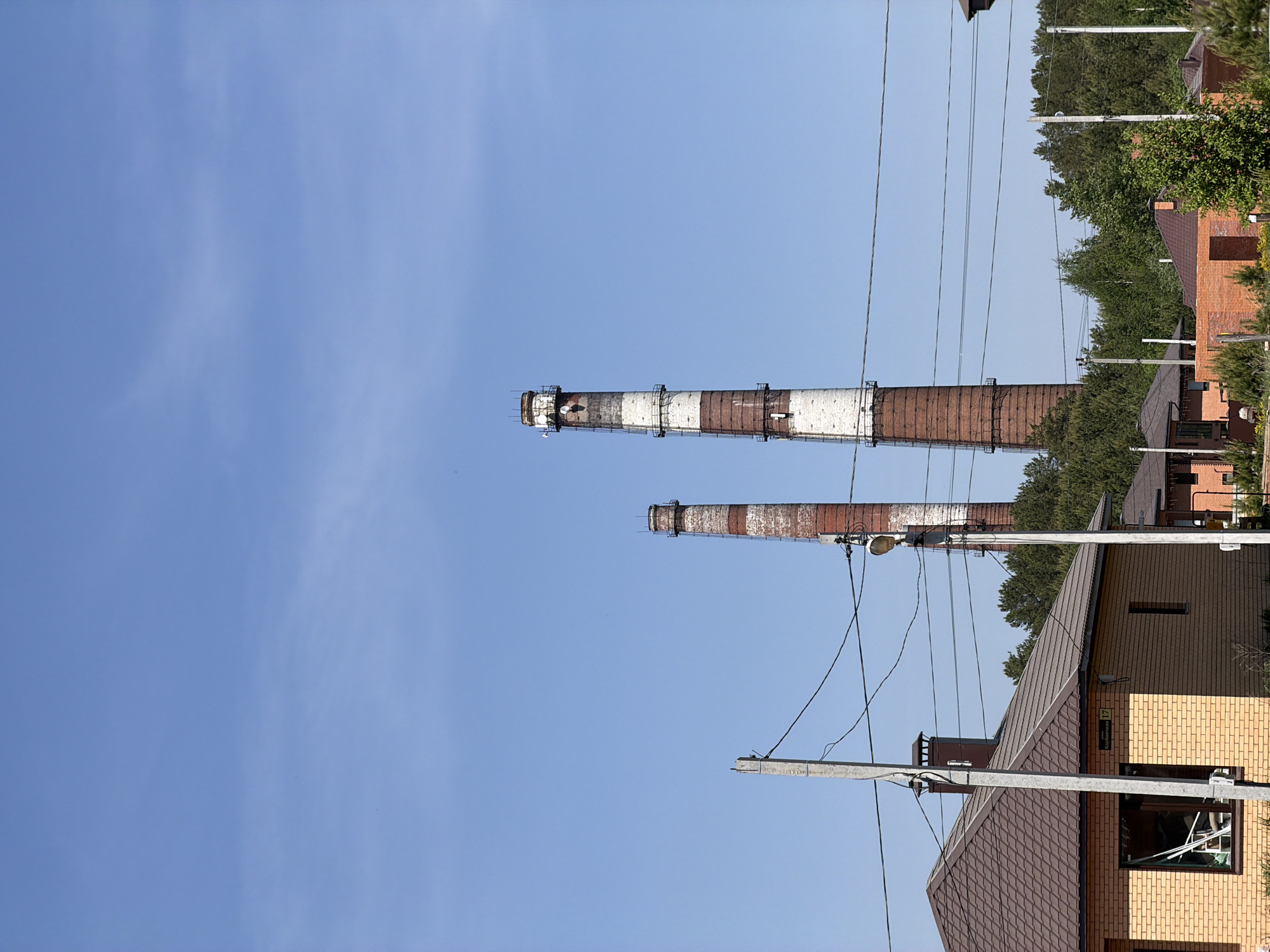 The image shows two tall, brick industrial chimneys against a clear, blue sky. The chimneys are banded with alternating red and white sections. One chimney appears taller than the other. Power lines and utility poles are visible in the foreground, running diagonally across the frame. The roofs and facades of residential buildings constructed with red and yellow bricks are partially visible on the right side of the image. The foliage from trees partially obscures some of the buildings. The location is identified as Chaikovsky, Russia.