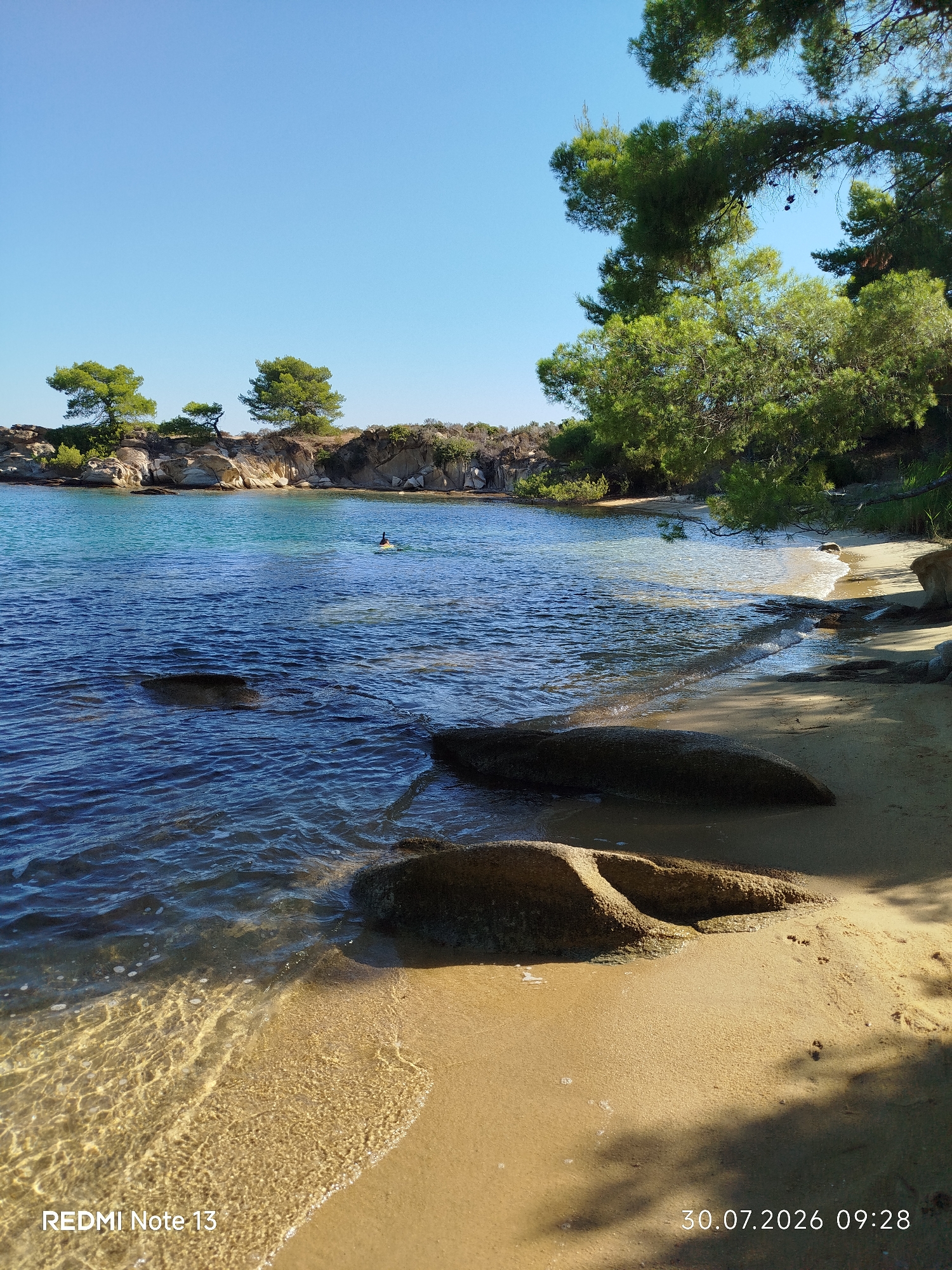 The image depicts a tranquil beach scene in Sithonia Municipal Unit, Greece, on July 30, 2026, at 09:28. The foreground is dominated by a sandy beach with several large, weathered rocks scattered across it, some partially submerged in the clear, shallow water. The water itself is a vibrant blue, reflecting the clear, cloudless sky above. Gentle waves lap at the shore, creating a foamy edge on the sand.
In the middle ground, the water deepens and the seabed becomes less visible, though some submerged rocks are still apparent. A person is visible in the distance, swimming or floating on a yellow inflatable object. The shoreline beyond the water features a rocky outcrop covered with sparse vegetation and pine trees. The trees, with their distinctive needle-like foliage, are a prominent feature, with some branches extending over the water and casting shadows on the beach. The overall impression is one of a peaceful, sunny day at a secluded coastal location. The bottom left corner of the image displays "REDMI Note 13" indicating the device used to capture the photo.