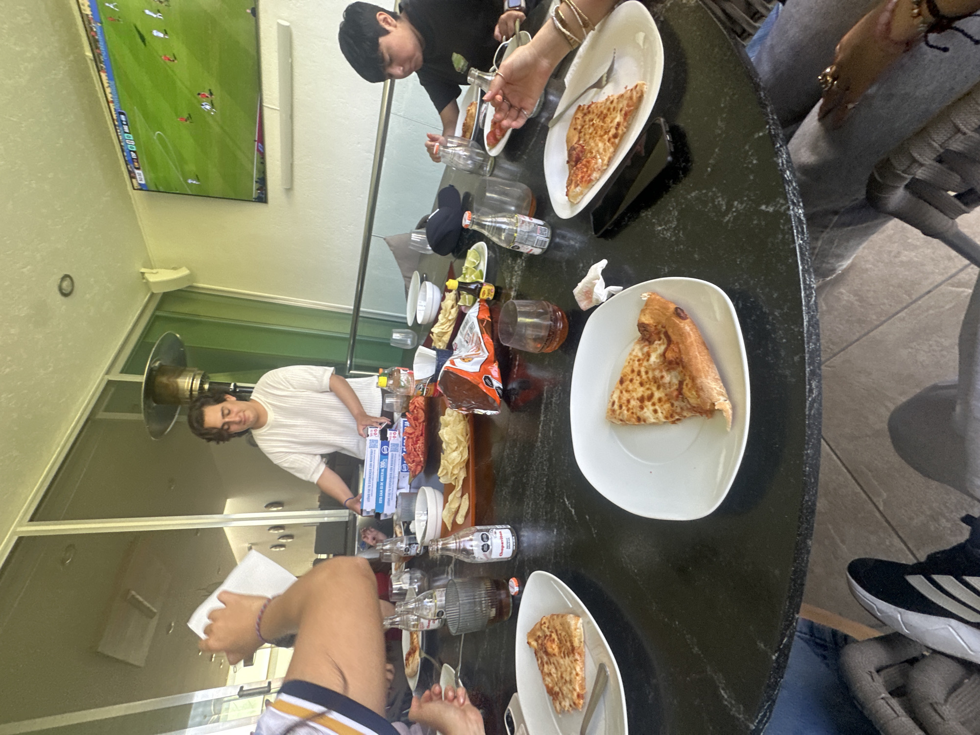 In San Bernardino Tlaxcalancingo, Mexico, a group of people are gathered around a large, black, circular table, enjoying a meal. The scene appears to be an outdoor patio or balcony, with large windows in the background revealing a glimpse of an interior space and a high-end patio heater. A large flat-screen television mounted on the wall displays a soccer match, suggesting a communal viewing experience. The table is laden with food, including several slices of pizza on white plates, bags of chips, lime wedges, and small bottles of what appear to be beverages. A young man in a white, short-sleeved shirt stands behind the table, holding a stack of what looks like pizza boxes, indicating he may be serving the food. Several individuals are seated around the table, with their hands visible, some reaching for food or drinks. The overall atmosphere suggests a casual, social gathering centered around food and entertainment.