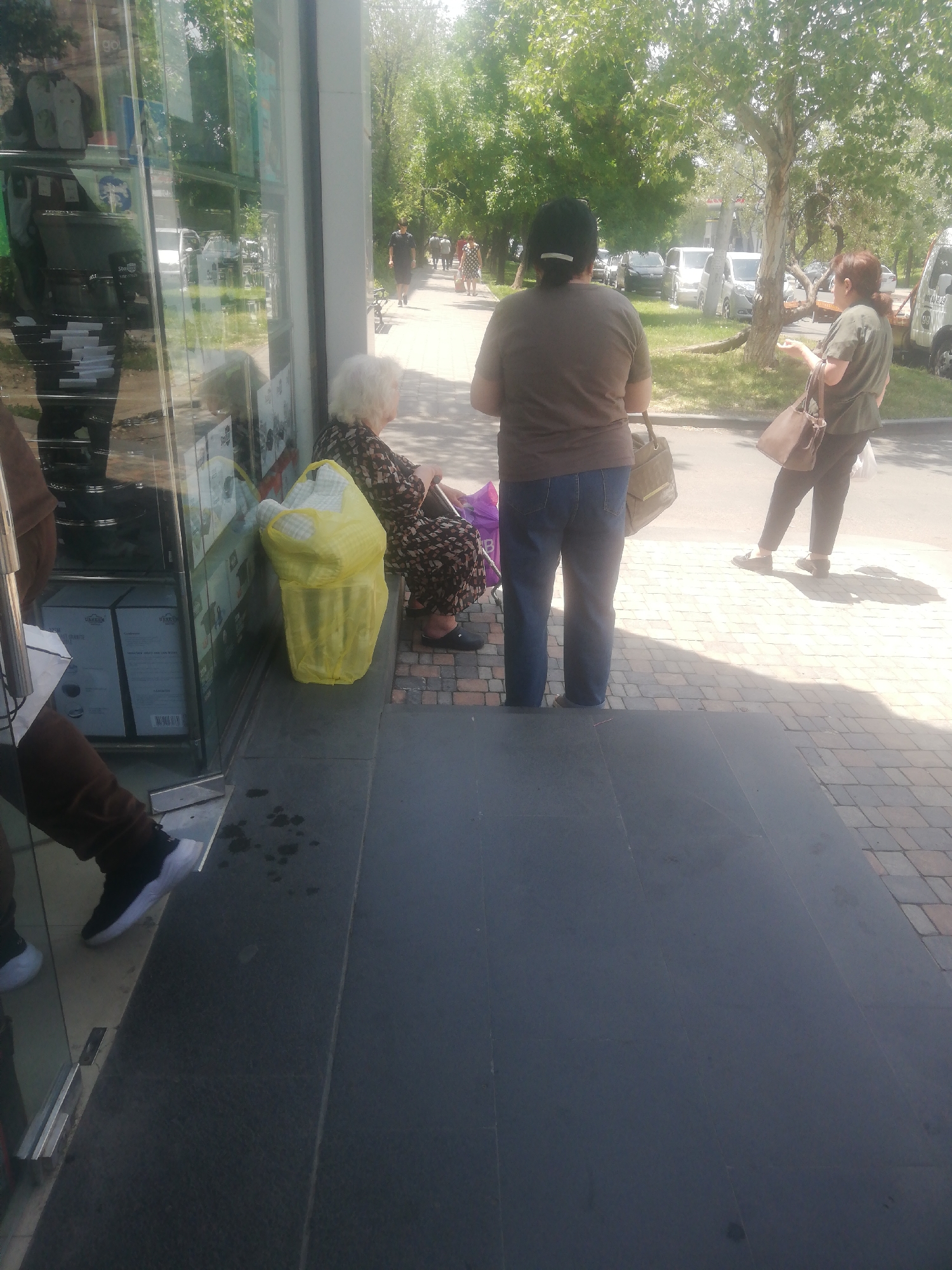 The image depicts a street scene in Yerevan, Armenia, on a sunny day. In the foreground, to the left, a person's legs and feet are visible, wearing dark brown pants and black and white sneakers. They are standing on a dark gray tiled surface. To the right of this person, an elderly woman with white hair is seated on a bench, wearing a dark patterned dress and dark slip-on shoes. She is holding a purple object and has a large yellow bag next to her. Behind her, a woman with dark hair pulled back is standing, wearing a dark gray t-shirt and blue jeans, and carrying a large tote bag. Further in the background, another woman in a green shirt and dark pants is walking away, carrying a brown shoulder bag and a white plastic bag. The scene includes a glass storefront reflecting trees and a street with several parked cars. The overall environment suggests a commercial area with pedestrian activity.