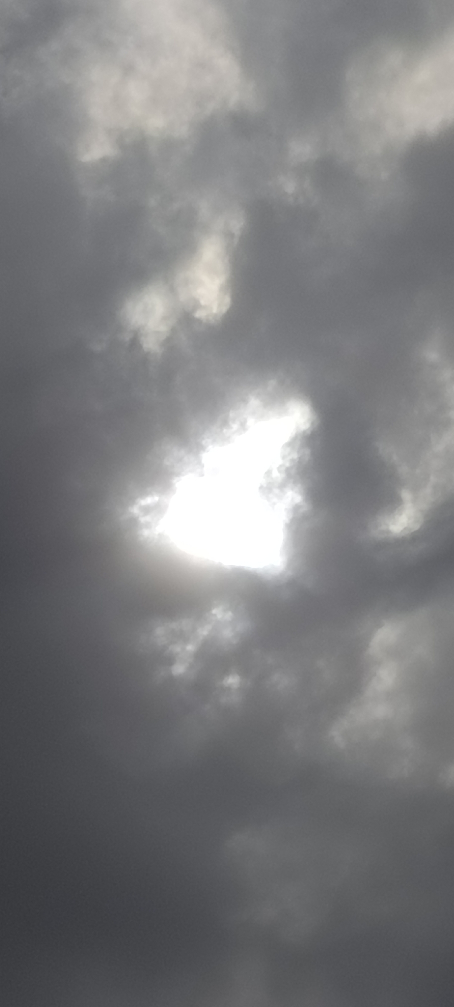 The image captures a sky dominated by dense, grey clouds, characteristic of an overcast or potentially stormy day. A bright, intensely luminous white area, likely the sun, is visible in the upper-middle section, attempting to penetrate the thick cloud cover. This bright spot creates a strong contrast with the surrounding darker, textured clouds, which vary in shades of grey, from deep charcoal to lighter, diffused tones. The overall scene focuses entirely on the atmospheric conditions, showcasing the interplay of light and shadow within the cloud formations. There are no people, objects, or visible text within the frame. The image conveys the appearance of daytime under heavy cloud cover.