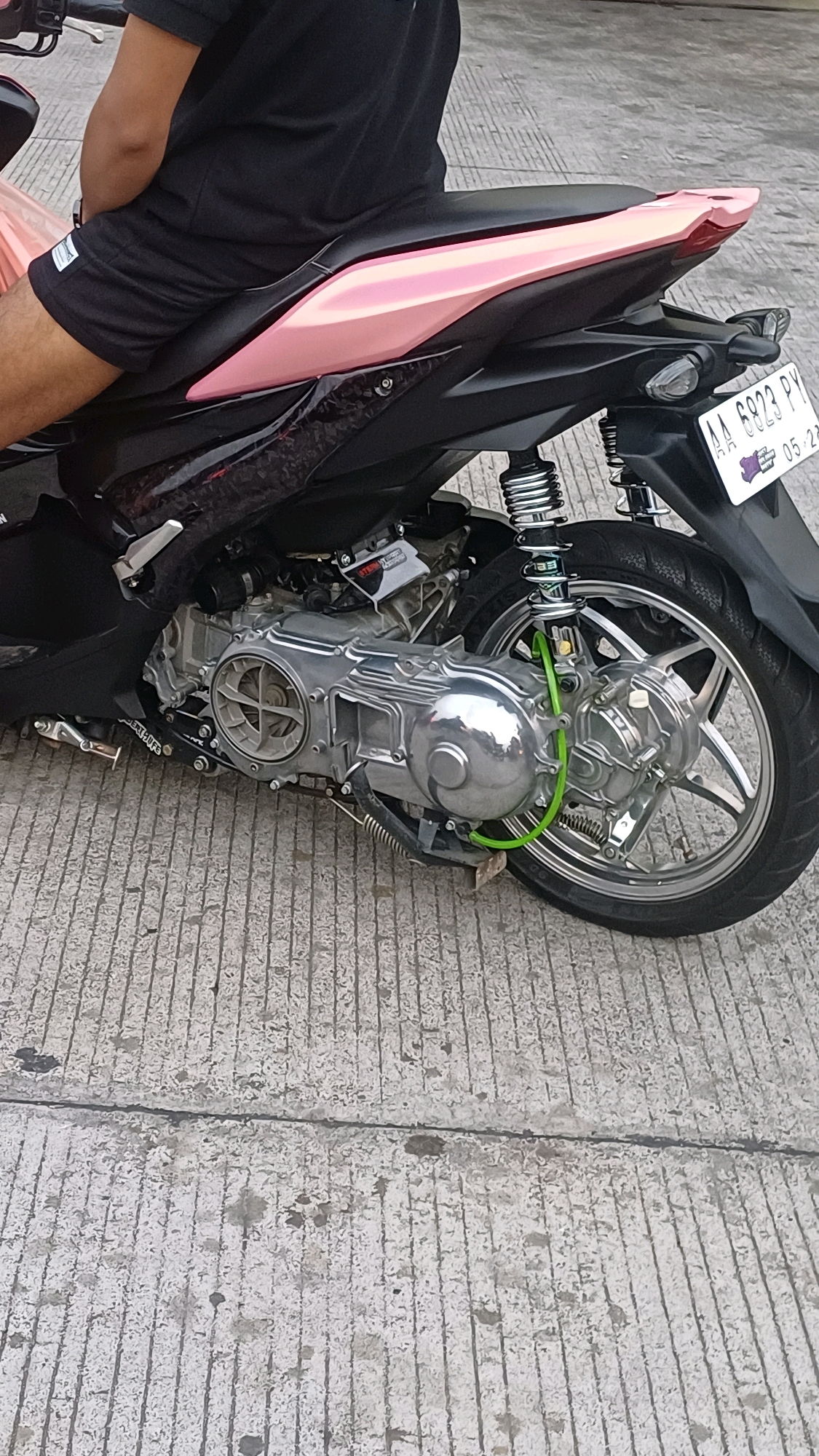 A person, wearing a black t-shirt and black shorts, is sitting on a black motorcycle with a pink seat cowl and pink body panels. The motorcycle is parked on a concrete surface with visible lines and some debris. A white license plate, reading "AA 6823 PY" and "05.28", is attached to the rear of the motorcycle. The scene appears to be outdoors, possibly on a street or parking area, given the concrete ground and the presence of a motorcycle. The lighting suggests it is daytime.