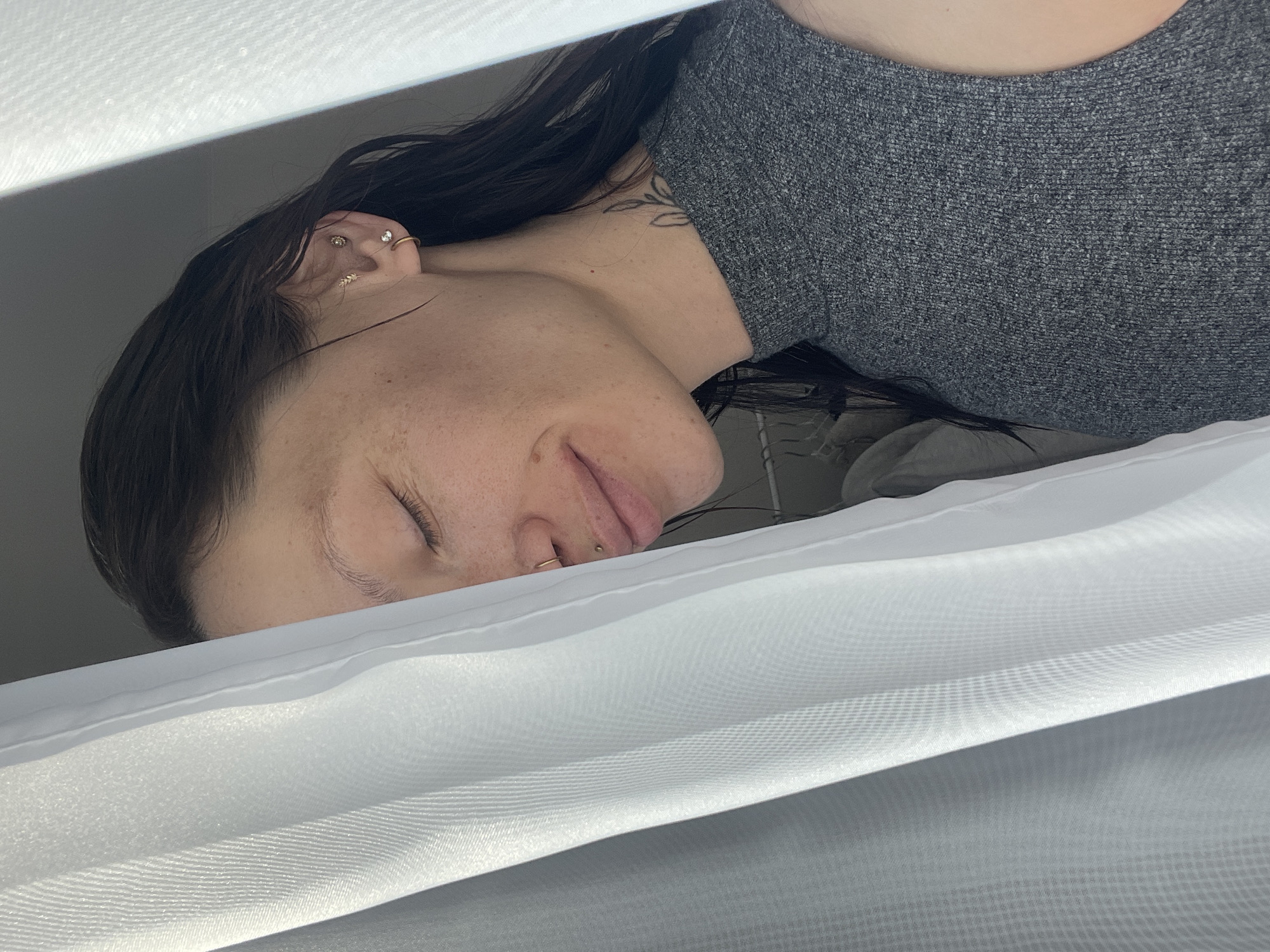 An intimate, angled close-up captures a young woman resting or sleeping, framed between layers of white, sheer fabric. Her head is tilted, with dark, long hair cascading around her face and neck. Her eyes are closed, and her expression is peaceful and serene, suggesting deep relaxation.
Notable details include multiple piercings on her visible left ear, featuring a mix of studs and a small hoop. She also has a small gold stud above her upper lip (philtrum piercing) and a gold hoop nose ring on her left nostril. A delicate, black-ink tattoo, possibly a botanical design, is partially visible on her neck/shoulder area, peeking out from under her dark hair. Her skin shows natural freckles across her cheek and forehead.
She is wearing a heather grey, possibly knitted or textured, top with a rounded neckline. The white fabric in the foreground and background creates soft, diffused lighting, suggesting an indoor setting, possibly with natural light filtering through curtains or bedding. The overall impression is one of quietude and calm. No text is visible in the image.
