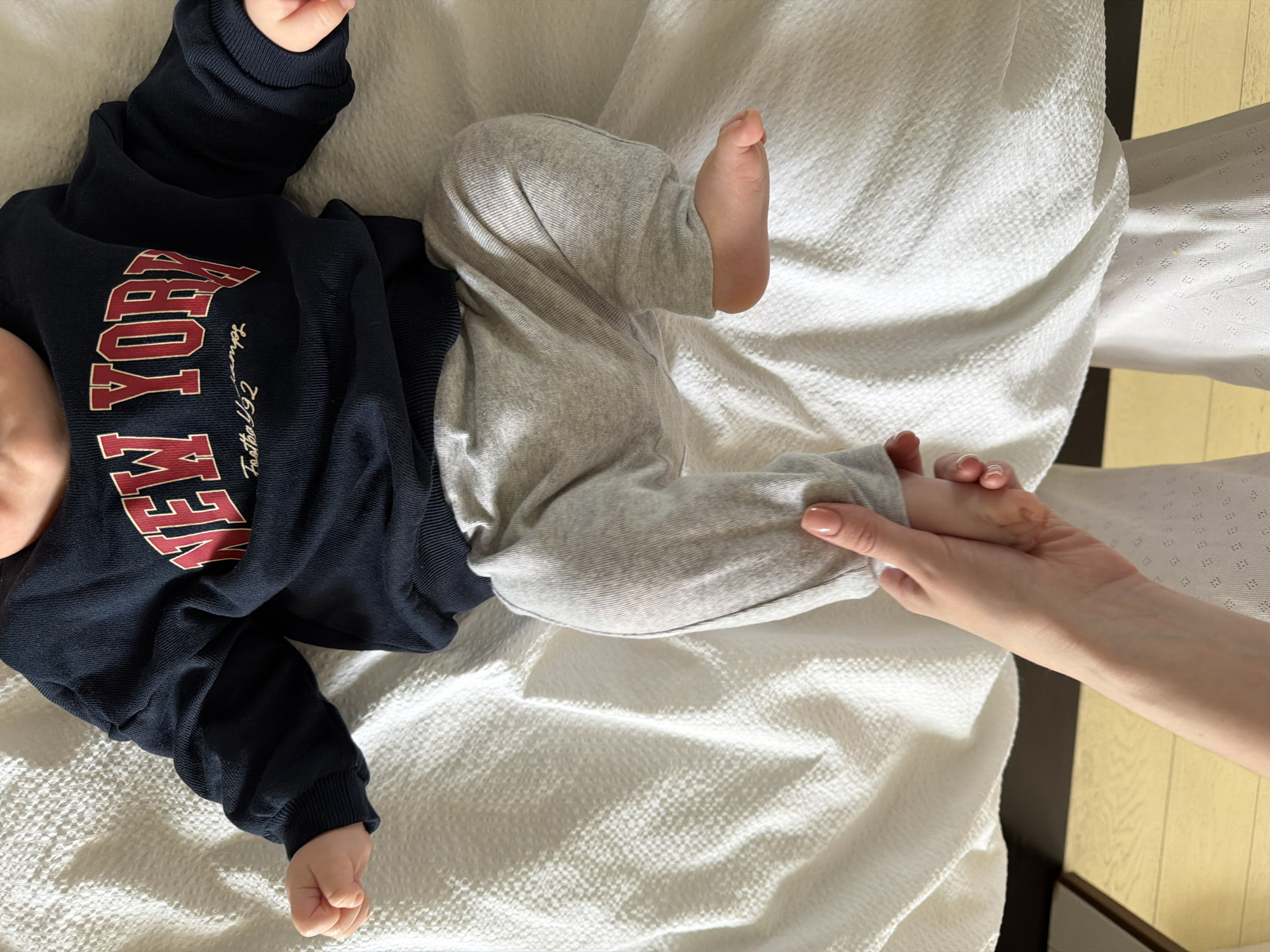 The image captures an overhead view of a baby lying on a white, textured blanket or bedding. The baby is dressed in a dark blue sweatshirt and light gray sweatpants. On the sweatshirt, the prominent text "NEW YORK" is displayed in red capital letters with a white outline, followed by smaller script text that reads "For the 192". An adult's hand, positioned on the right side of the frame, is gently holding one of the baby's feet, suggesting a moment of affectionate care or interaction. The baby's body is oriented diagonally across the frame, with only a part of their head visible at the top left. The background reveals an indoor setting, likely a home in Khimki, Russia, with a dark surface and light wooden flooring visible to the right. The overall scene conveys a sense of tenderness and calm.