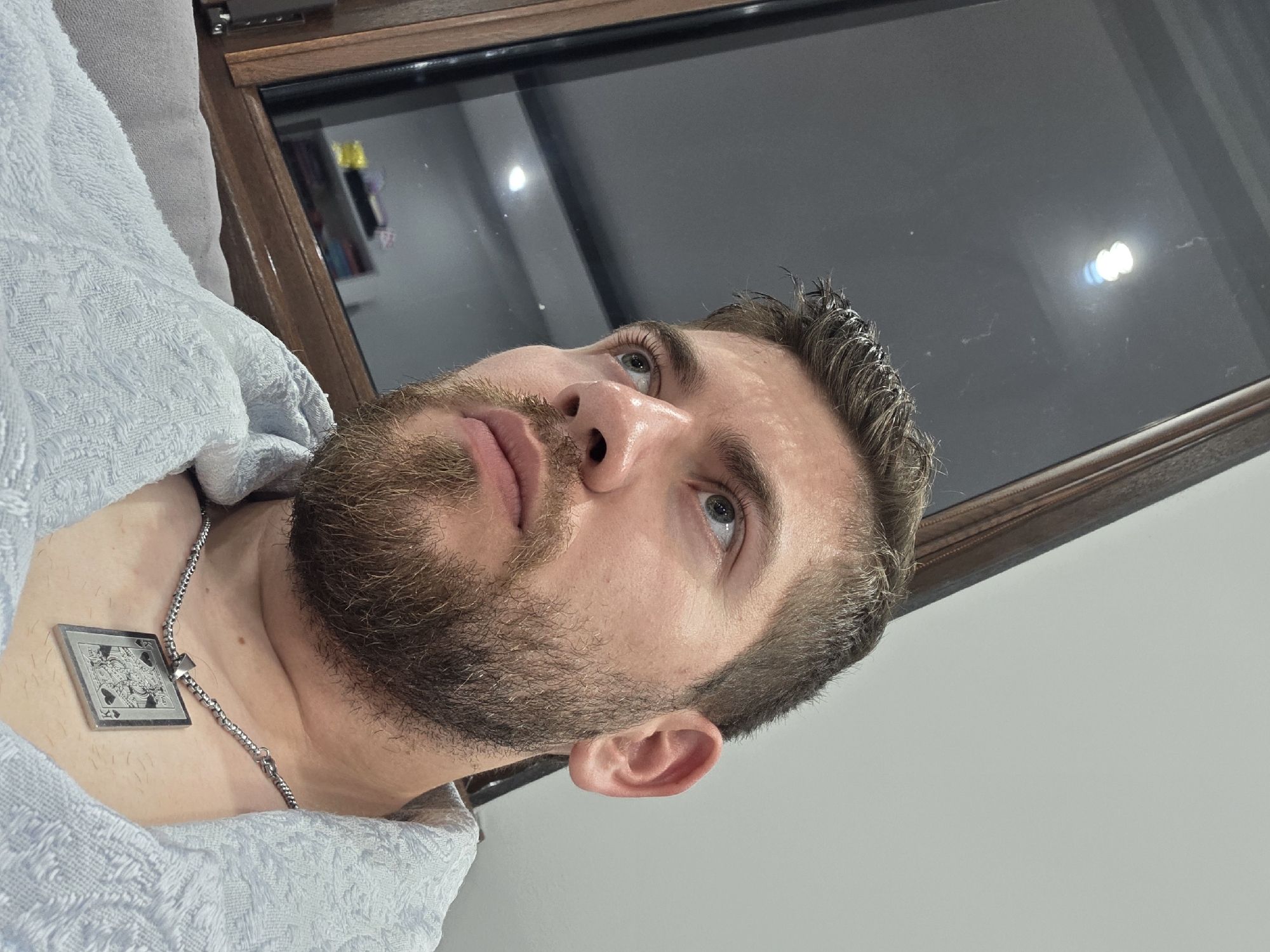 A close-up, slightly angled, medium shot features a man with a beard, fair skin, and short brown hair. He wears a silver chain necklace with a square pendant depicting a card suite, likely a spade or club. He appears to be wrapped in a light blue towel or robe. The background includes a dark framed window or mirror reflecting light and possibly other objects. The scene appears to be indoors, potentially a bathroom. There is no information to associate the location with Yagodovo, Bulgaria.