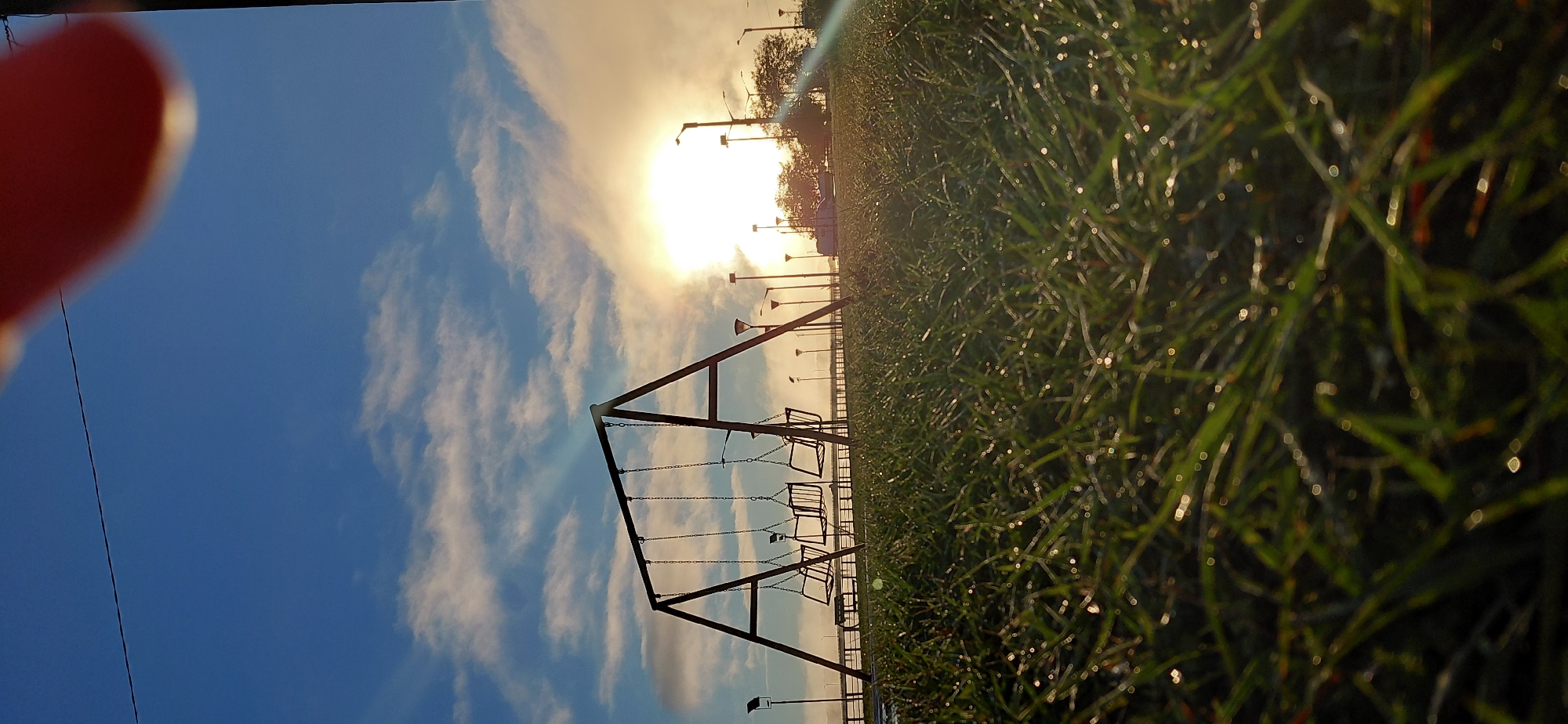 The image captures a low-angle view of a playground swing set in the early morning light, set against a backdrop of a bright sun partially obscured by clouds and a clear blue sky. The foreground is dominated by dewy green grass, with individual blades and water droplets sharply in focus, creating a bokeh effect. Behind the grass, a metal A-frame swing set holds three empty swings. Beyond the swing set, a paved walkway or road is visible, lined with streetlights. In the distance, trees and vehicles are silhouetted against the sun, which casts a warm glow, creating a slightly hazy atmosphere. The scene suggests a tranquil park setting, likely in Encarnacion, Paraguay, given the presence of the distinctive lampposts seen in similar public spaces along the Costanera. There are no people visible in the frame.