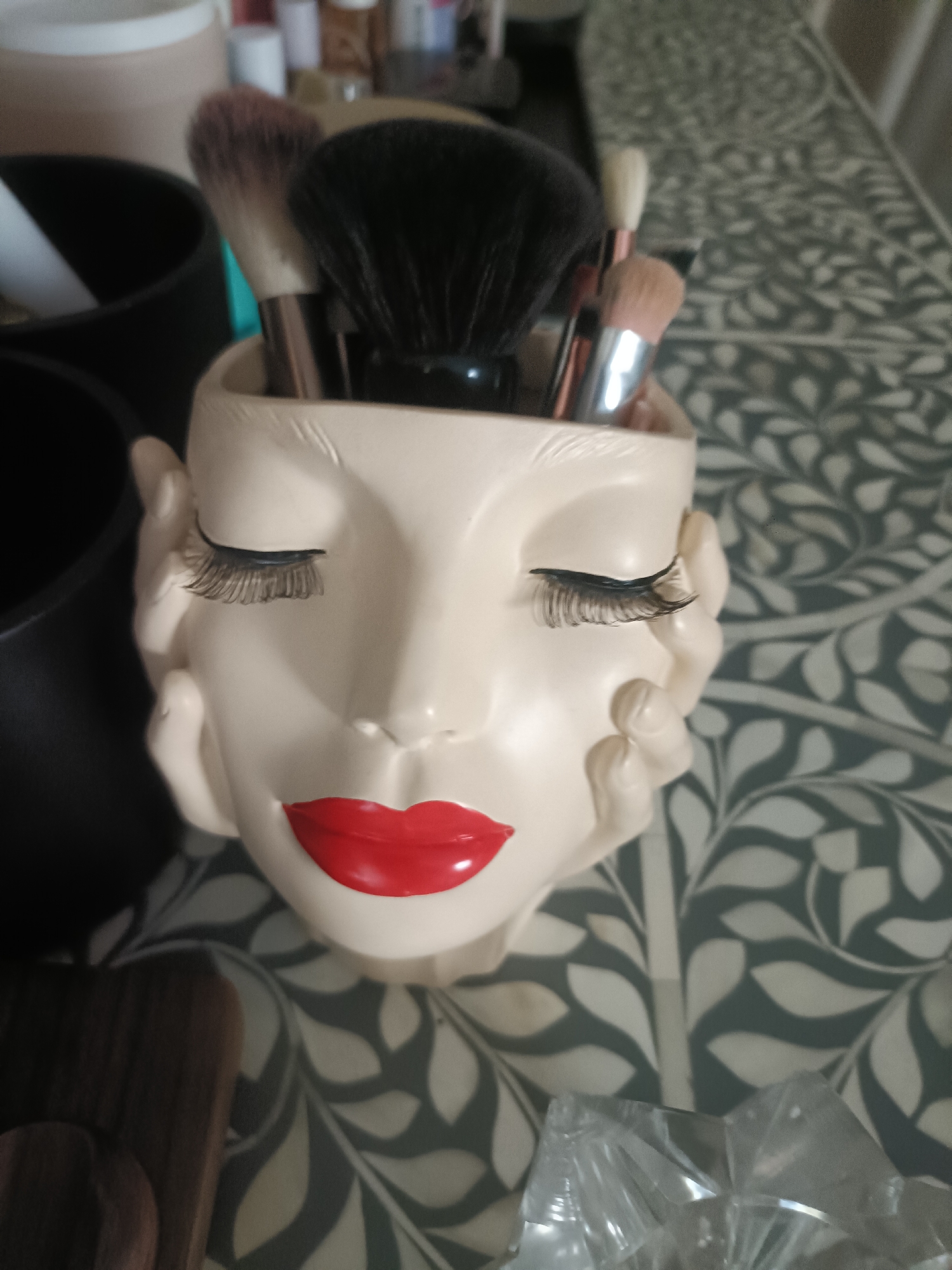 A close-up shot focuses on a ceramic holder shaped like a woman's face, filled with makeup brushes. The holder is cream-colored with bright red lips and has applied false eyelashes. The makeup brushes have black and brown bristles and rose-gold metallic handles. The holder is placed on a patterned tablecloth with grey and white floral designs. To the left of the holder, a black container and a portion of another cream-colored container are visible. To the right, a clear plastic object, possibly a crystal vase or decorative item, is partially visible. The scene appears to be indoors, likely on a table. No specific location in Dubai, United Arab Emirates, can be identified from the image.