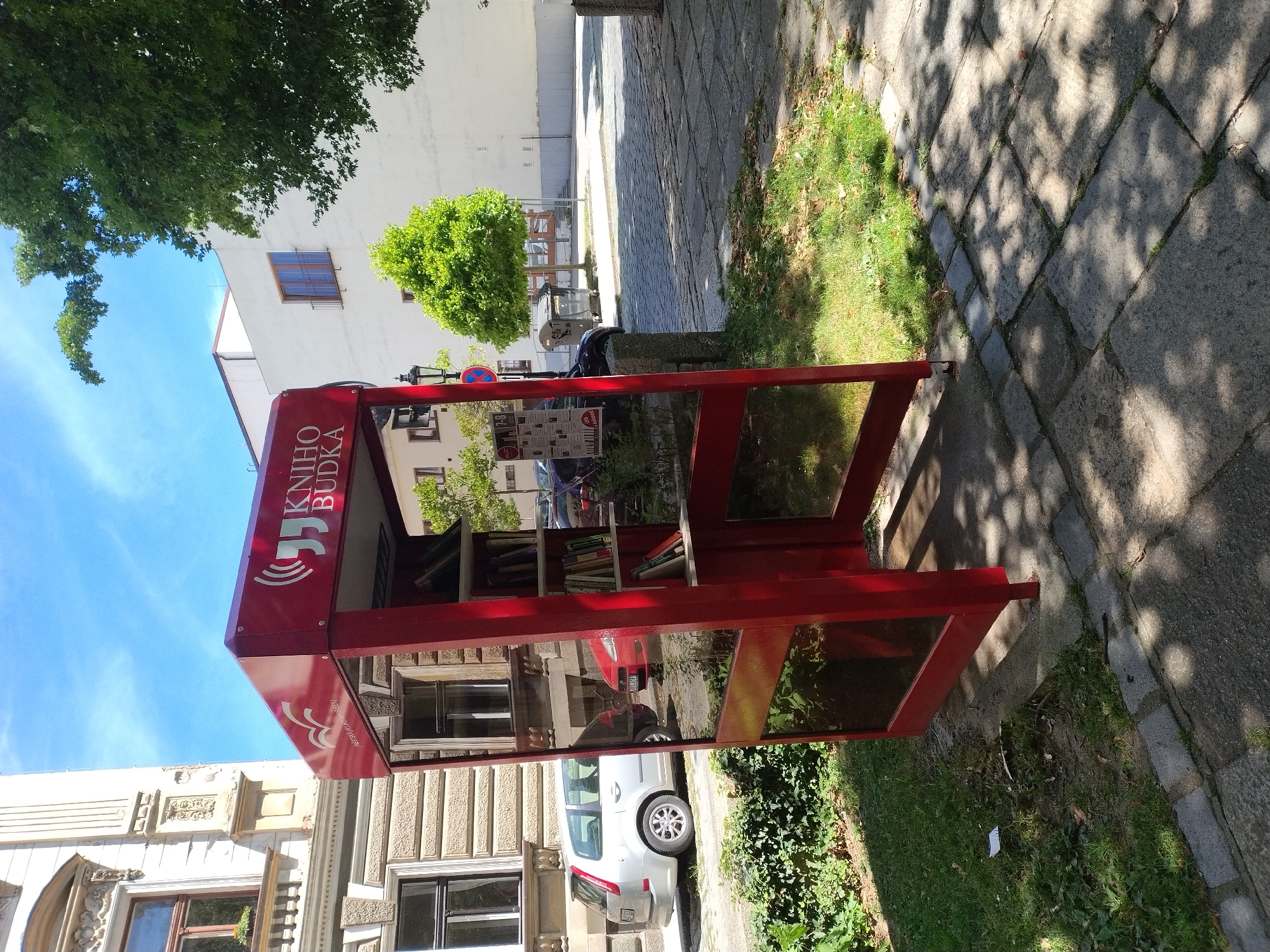 An outdoor, repurposed red telephone booth serves as a public book-sharing station in Písek, Czechia. The booth is situated on a paved area beside a patch of grass and a tree-lined street. Inside, several shelves display a collection of books. In the background, a silver car and a red car are parked on the street, with buildings and trees visible behind them. The sky is blue with scattered clouds, suggesting a sunny day. The scene appears to be a public space, possibly a street or park, in the city.