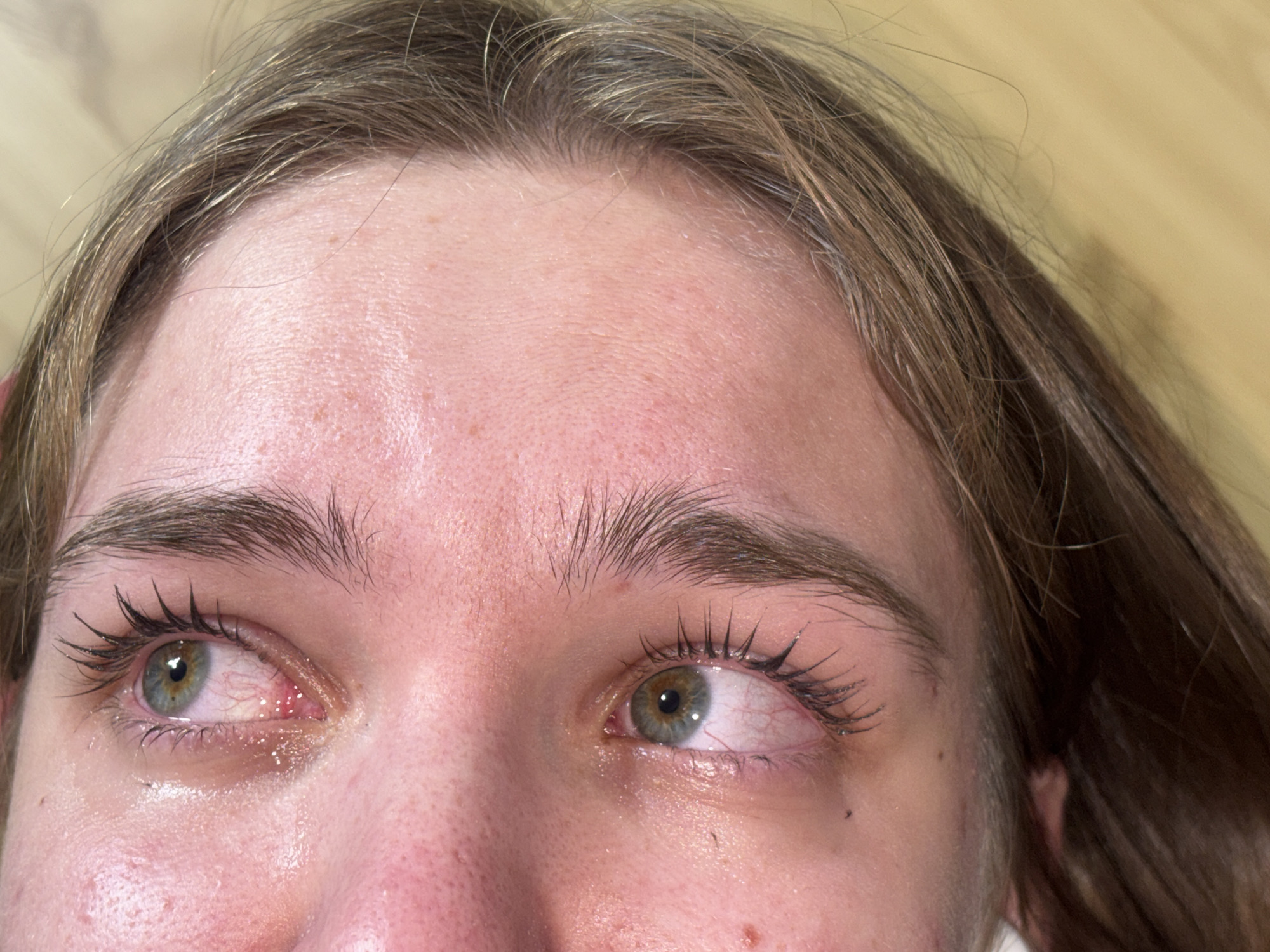 A close-up shot of a person's face, focusing on the eyes, forehead, and eyebrows. The person has light brown hair, and their eyes are hazel with prominent eyelashes coated in mascara. The left eye appears bloodshot and watery, suggesting irritation or crying. The skin is fair, with visible pores and some redness. The overall lighting is bright and even. The background is out of focus, revealing a light-colored wooden surface. There is no information about location.