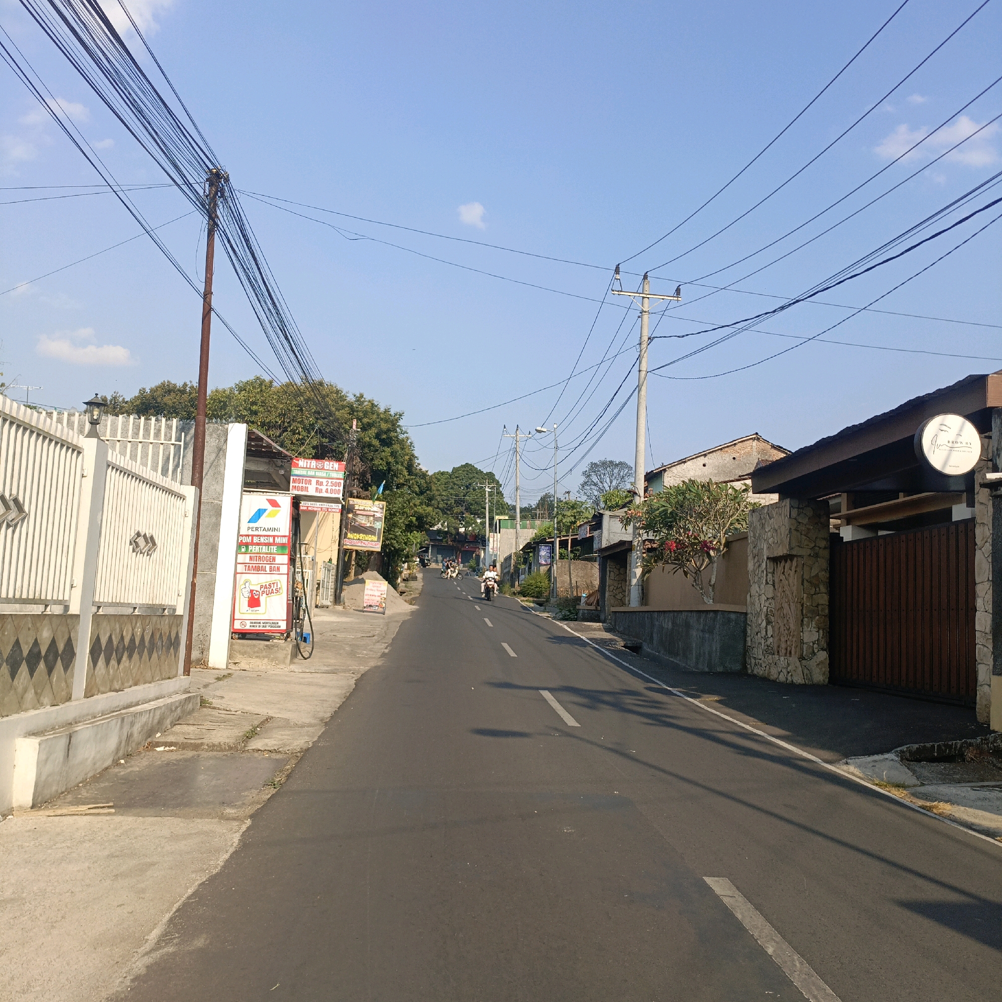 The image shows a street scene in Salatiga, Indonesia. The street is paved and lined with buildings on both sides. There are utility poles with numerous wires running across the sky. On the left side of the street, there is a white fence with a diamond pattern and a Pertamina gas station with advertising for fuel and tire services. A bicycle is parked near the gas pump. Further down the street, there are houses and trees. Two people are riding motorcycles on the street, and shadows of utility poles and possibly trees are cast on the road. The sky is clear blue with a few small white clouds. On the right side of the street, there is a building with a dark brown roof, stone walls, and a large wooden gate. A circular sign for "BROW RY" is visible above the gate. The overall impression is a typical street in a residential or commercial area in Indonesia.
