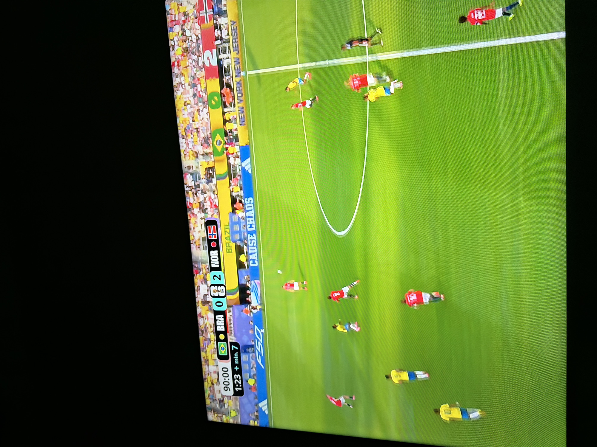 This is a photograph of a soccer match being shown on a television screen. The score displayed at the top of the screen indicates that Brazil (BRA) is losing to Norway (NOR) with a score of 0-2. The game time is 90:00 with an additional 1:23 minutes of stoppage time. The players on the field are wearing red and yellow jerseys, representing the two teams. The environment is a brightly lit soccer stadium with a large crowd in the background. The photograph is taken at an angle, suggesting it is a candid shot of someone watching the game on TV. The city of Ognianovo, Bulgaria is not depicted in this image.