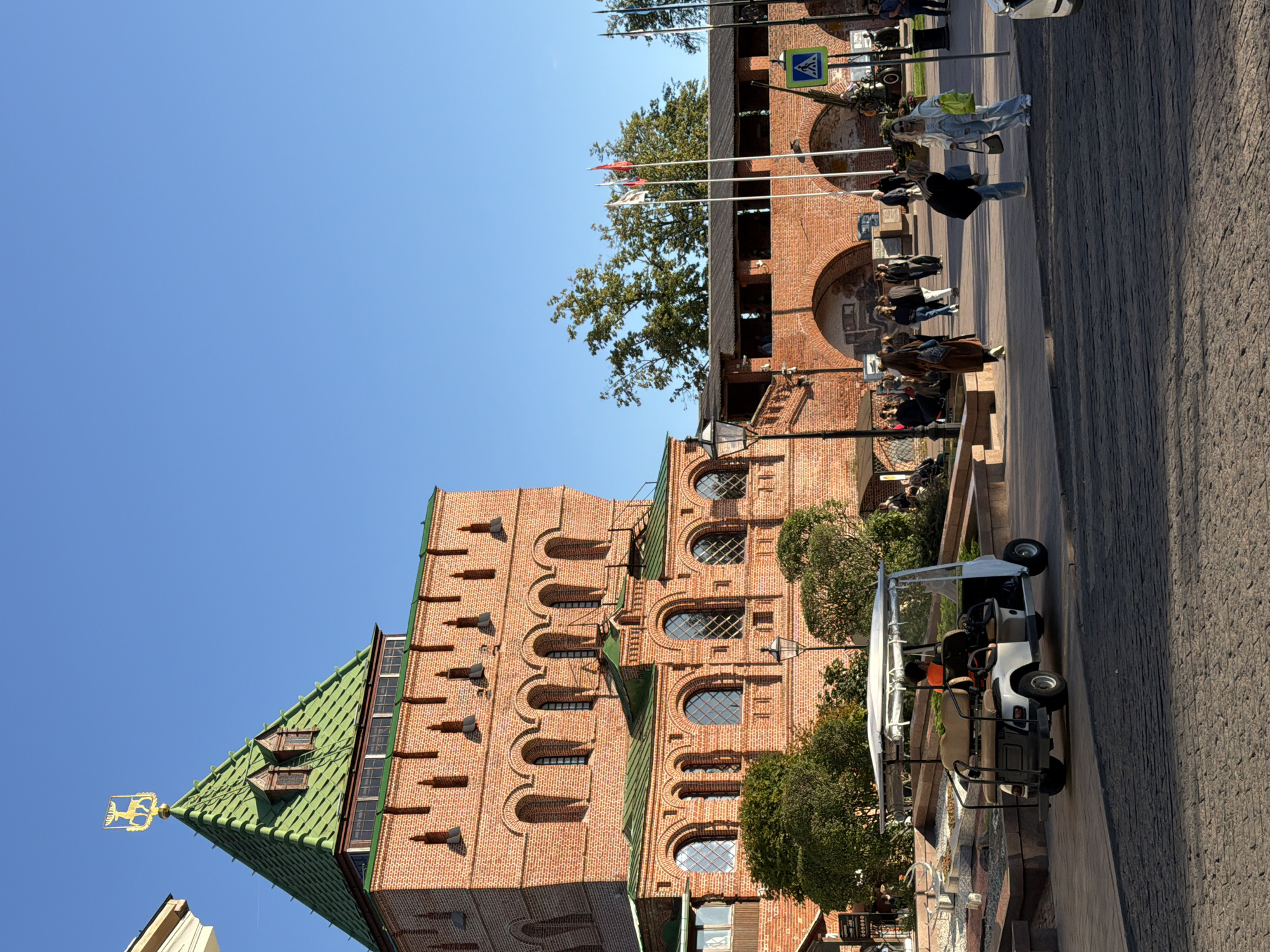 In Nizhny Novgorod, Russia, a golf cart with a single occupant drives across a cobblestone street in front of a large, brick fortress with a green, pyramid-shaped roof. The fortress has multiple arched windows and decorative brickwork. People are seen walking on the sidewalk near the fortress, some carrying bags. Flags are visible on poles in the background, and trees grow behind a section of the fortress wall. The sky is clear and blue, indicating a sunny day.