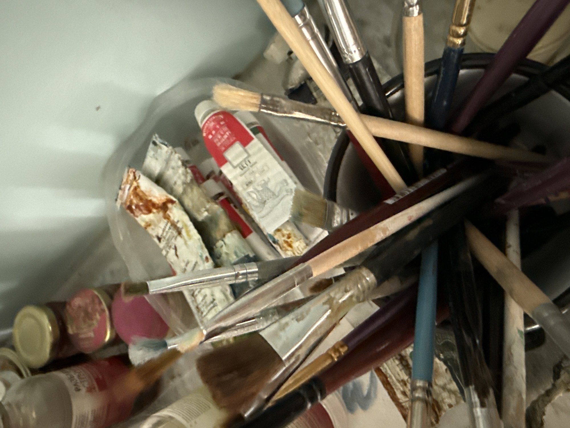 A close-up, slightly blurred, eye-level shot focuses on a collection of art supplies. The primary subjects are numerous paintbrushes, varying in size, bristle type, and handle color. The handles are a mix of wood tones, black, dark red, and a striking teal. Some brushes are clean, while others appear to be stained with paint. Interspersed among the brushes are tubes of paint, one prominently featuring a red and white design. The supplies are loosely contained within a bucket or container, though only the upper rim of this container is visible. In the background, hints of other items can be discerned, including what appear to be jars with metallic lids and a partially visible bottle with a red label, suggesting an art studio or workspace. The lighting is somewhat dim, casting shadows and contributing to the overall slightly cluttered and artistic atmosphere. There is no indication of people or any specific location within Vladivostok, Russia.