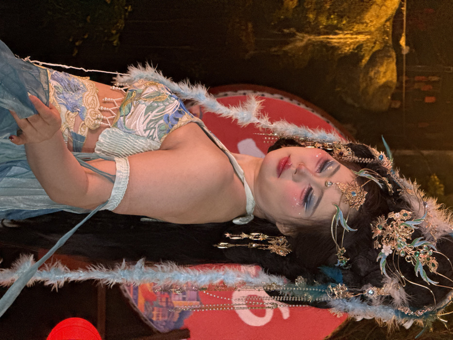The image features a person, likely female, lying down and elaborately dressed in a fantasy or traditional-inspired costume. She wears a light blue and gold top with intricate floral embroidery, pearl accents, and a soft, feathery blue trim. Her long, dark hair is adorned with numerous ornate golden hairpins, tassels, and blue-feathered accessories. Her makeup is dramatic, featuring blue eyeshadow with glitter, pink blush, and red lipstick, creating a serene expression with her eyes gently closed.
The person is positioned on a dark surface, with her head and hair resting on a large red circular object that bears partially visible white text, possibly "SUPREME." A red lantern or light source is partially visible in the bottom left corner. The background is dark and out of focus, suggesting a nighttime or evening setting, with blurred warm lights indicating a distant urban environment.
The activity appears to be a staged pose for a photograph, likely a costume shoot or artistic portrait. The overall aesthetic is highly stylized and ethereal, highlighting the intricate details of the attire and makeup. No specific location cues are visible within the image itself beyond the East Asian aesthetic of the costume.