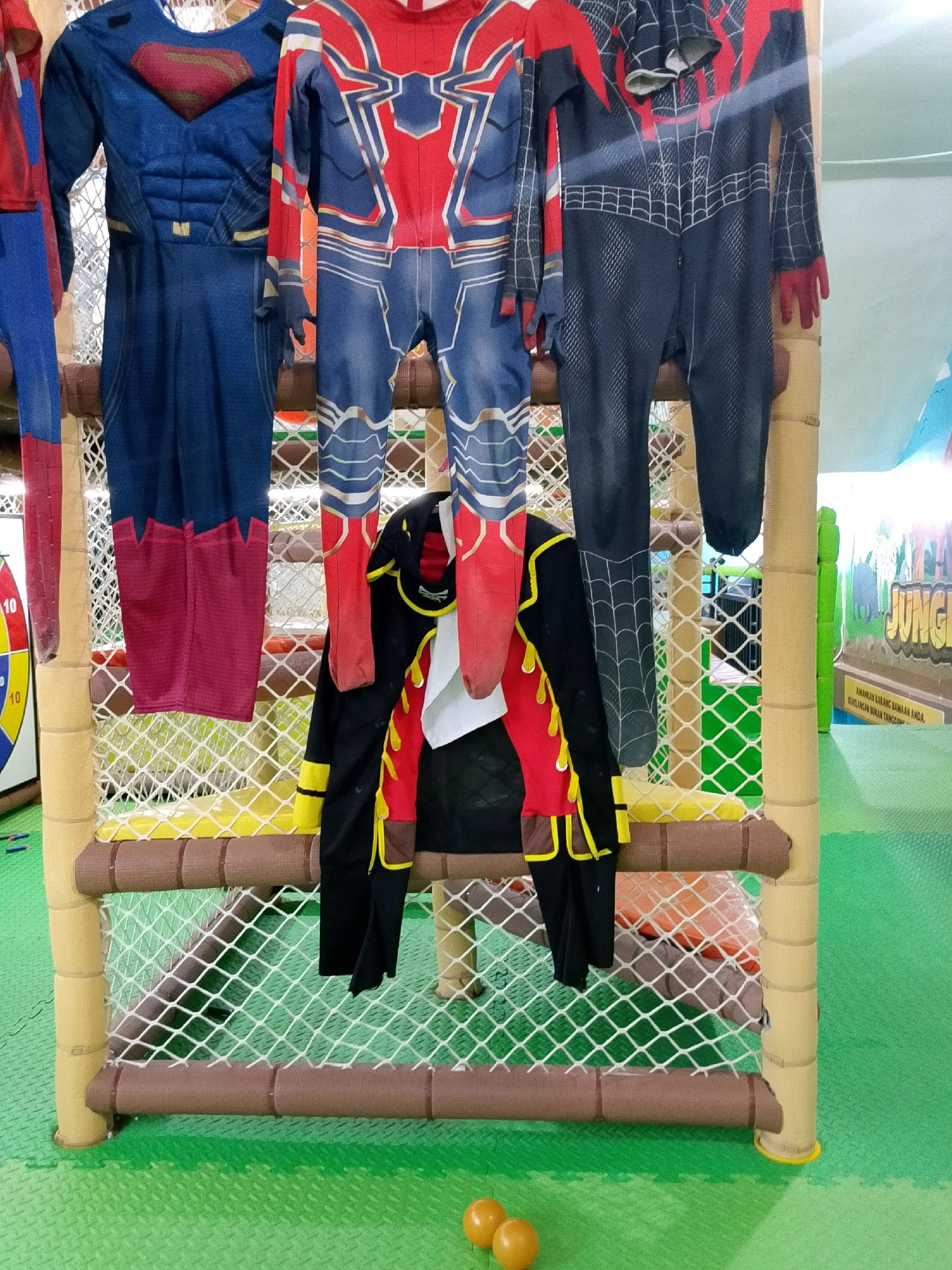 In a brightly lit indoor play area, possibly a children's amusement center in Banjarmasin, Indonesia, a collection of superhero costumes hangs on a netting structure. The costumes include a blue Superman suit with red and gold accents, an Iron Man suit with red, blue, and gold detailing, and a black and red Spider-Man suit. A black and yellow pirate-style jacket is also displayed. The play area features green padded flooring and brightly colored walls, one of which displays a jungle-themed mural with the word "JUNGLE" prominently featured. Two small orange balls are visible on the floor in the foreground. The overall impression is one of a fun and engaging environment for children.