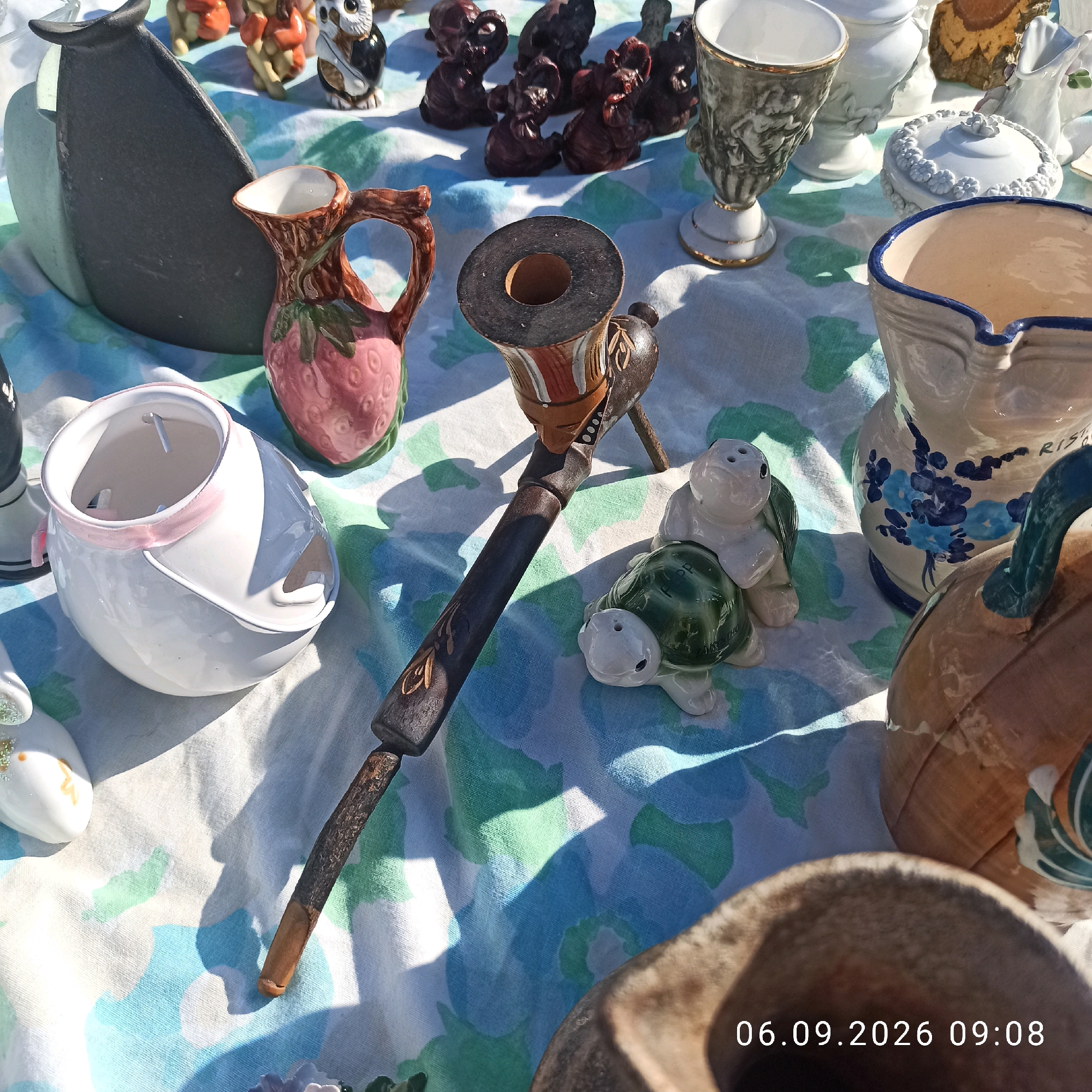 A flat lay photograph displays an assortment of ceramic and decorative objects spread across a blue and white patterned tablecloth. The items include a dark gray, irregularly shaped vase or vessel, a small pitcher shaped like a strawberry, a white ceramic bowl with a pink ribbon detail, a pipe-like object with a dark brown stem and a decorative mouthpiece, two ceramic turtle salt and pepper shakers, a white vase with blue floral designs, a white lidded container decorated with floral motifs, a tall, ornate gray vase with gold trim, and several small, dark-colored animal figurines. In the foreground, the edge of a large, brown, gourd-shaped ceramic object is visible. A timestamp "06.09.2026 09:08" is superimposed on the bottom right of the image. The scene is illuminated by bright sunlight, casting sharp shadows across the tablecloth. The arrangement suggests a stall or display, possibly at an outdoor market or fair. The specific location of Sannicandro di Bari, Italy, is not visually present within the image itself.