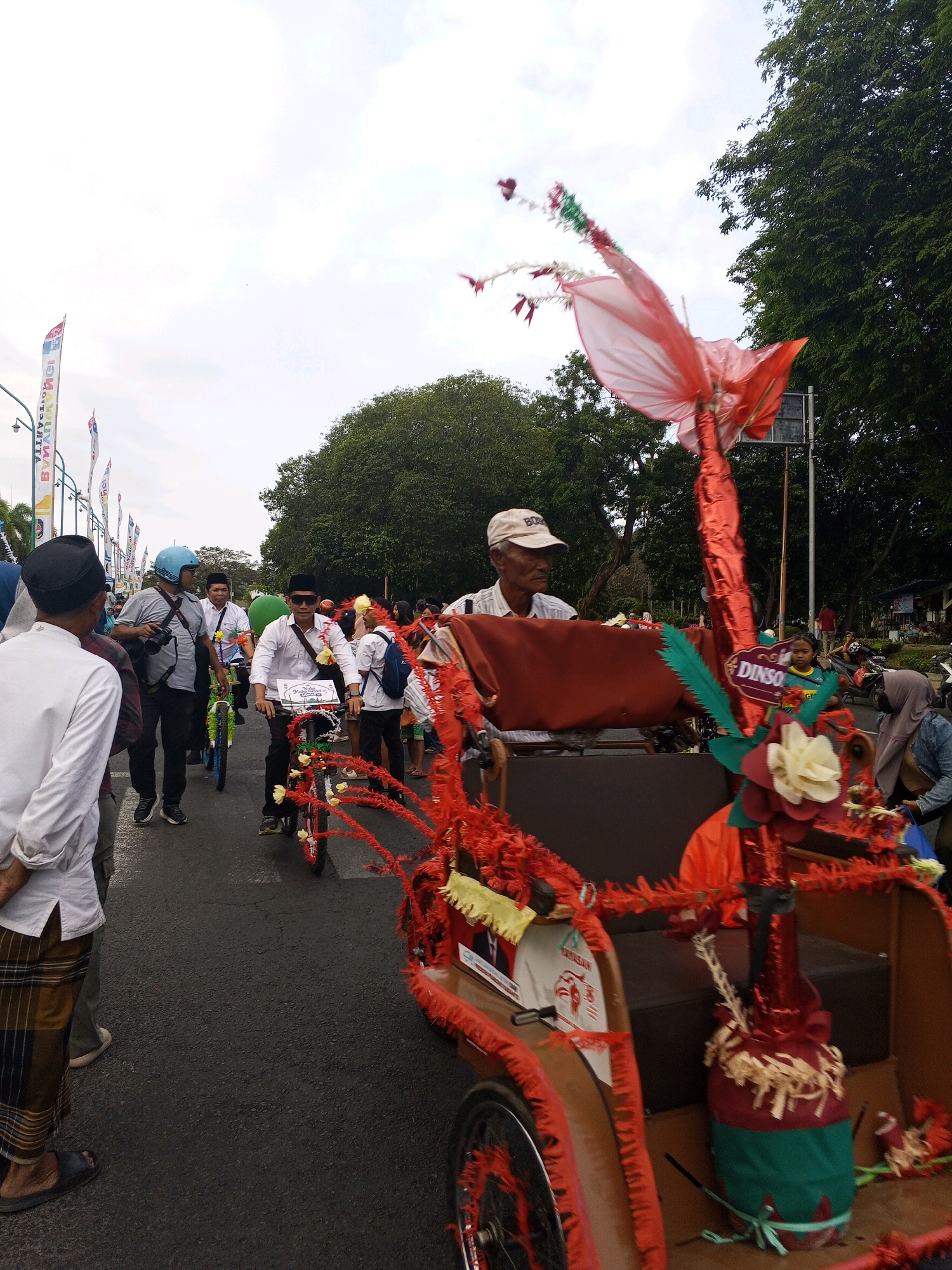 In Banyuwangi, Indonesia, a festive procession is underway on a paved road under a cloudy sky. The foreground features a vibrantly decorated becak, a pedal-powered tricycle, adorned with red tinsel, fabric flowers, and a tall, red, kite-like ornament. A man in a white cap and shirt sits in the becak. To the left of the becak, a man in a white shirt and black peci hat, wearing sunglasses and a garland, rides a bicycle decorated with red and yellow streamers. Behind him, another man in a blue helmet, also wearing a garland, walks and holds a camera. Further back, more people on bicycles and walking can be seen in the procession. Banners with colorful text are visible along the roadside, and lush green trees line the street on the right. In the background, other vehicles and pedestrians are present, suggesting a public event or parade.