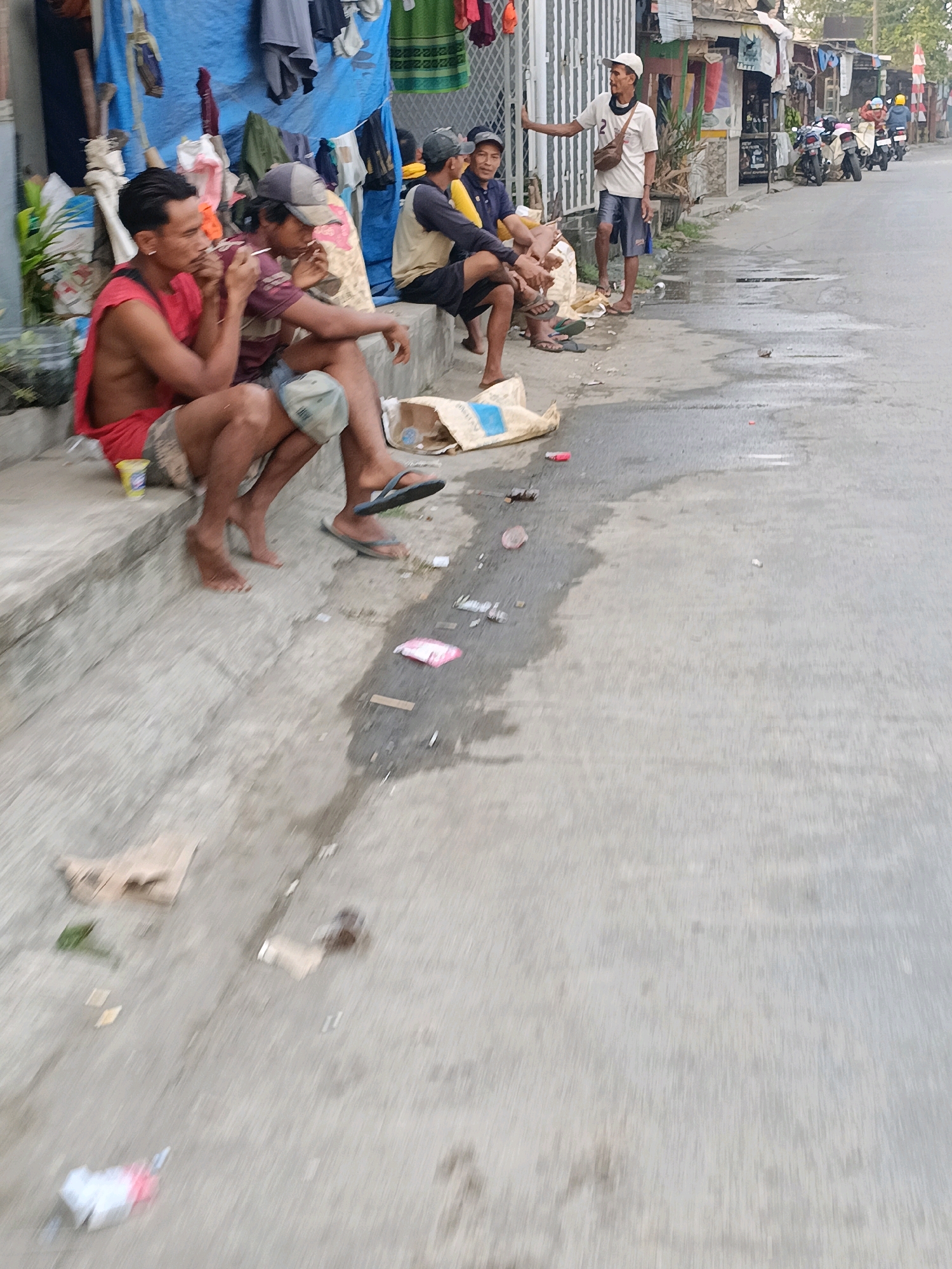 The image depicts a street scene in Tangerang, Indonesia. In the foreground, four men are seated on a concrete ledge. The man on the far left, wearing a red tank top and patterned shorts, is smoking. Next to him, a man in a burgundy shirt and denim shorts also appears to be smoking. Further to the right, two other men are seated, one wearing a yellow shirt and black shorts, and the other in a darker shirt and shorts.
Behind the seated men, a blue tarp hangs from a structure, with various items of clothing and bags suspended from it. A white metal fence borders the street to the right. Several motorcycles are parked in the background, and a few individuals are standing near the fence. The street itself appears wet in places, with scattered litter visible on the ground, including plastic bags, paper wrappers, and other debris. The overall impression is one of a casual gathering in a somewhat cluttered urban environment.