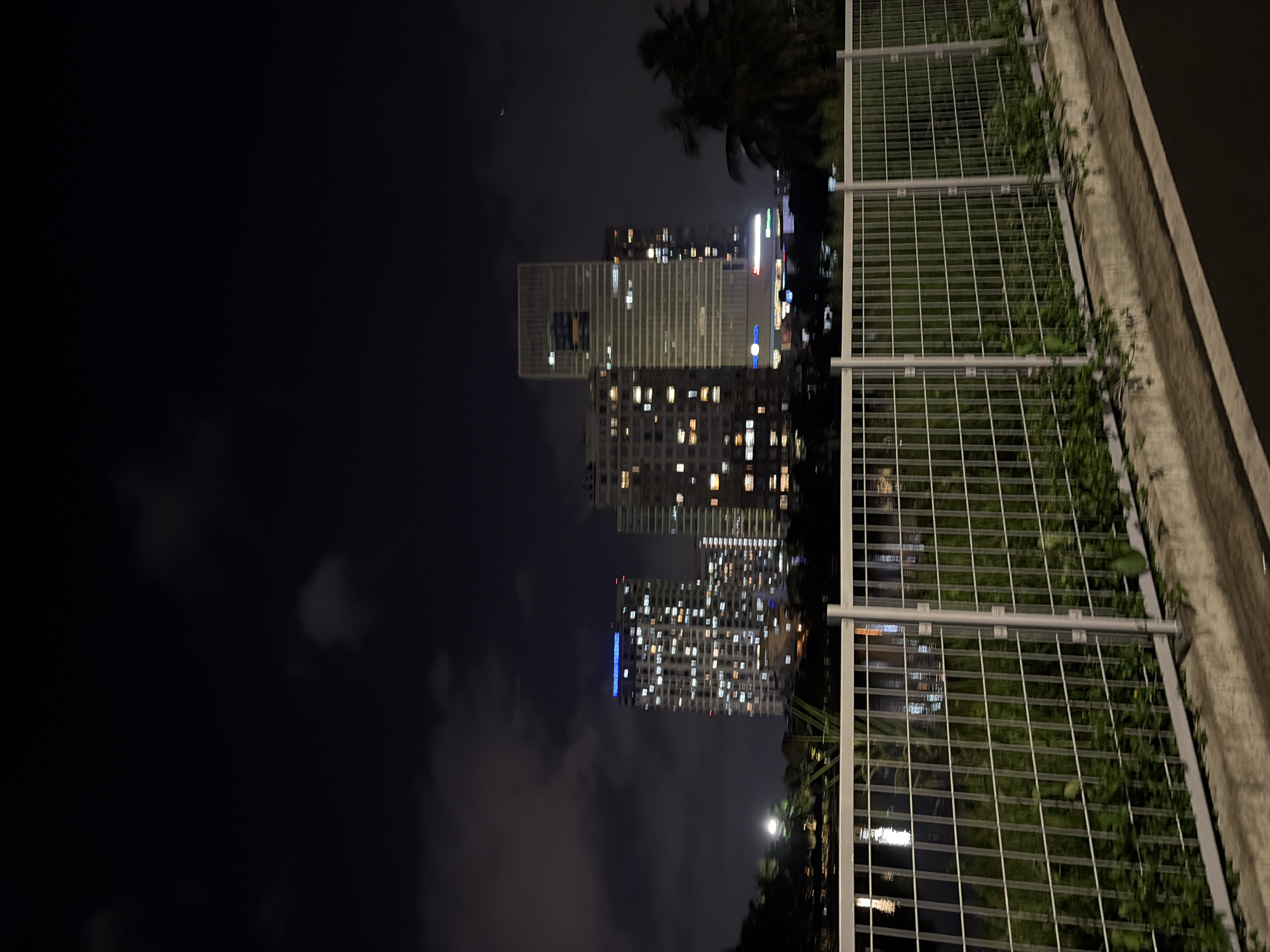 The media file presents a nighttime cityscape in Thủ Đức, Vietnam, captured from an elevated perspective. In the immediate foreground, a concrete wall or ledge runs horizontally, topped by a white metal railing with a grid pattern. Green vines and foliage are seen growing along this railing and spilling over the wall.
Beyond the foreground structure, several modern high-rise buildings dominate the mid-ground. Many windows across these multi-story structures are illuminated, casting warm glows and indicating human activity within. One notable building features a distinctive large square cutout near its upper section. Dark silhouettes of palm trees are interspersed among the buildings, hinting at a tropical climate. The sky above is dark, characteristic of night, with scattered clouds partially obscuring the view and creating a soft haze. A faint crescent moon is visible in the upper right quadrant of the sky.
The overall scene conveys the quiet yet active ambiance of a city after dark. There are no people visible, and no specific events are overtly taking place, but the numerous lit windows suggest a bustling urban environment. No discernible text is visible anywhere in the image.
