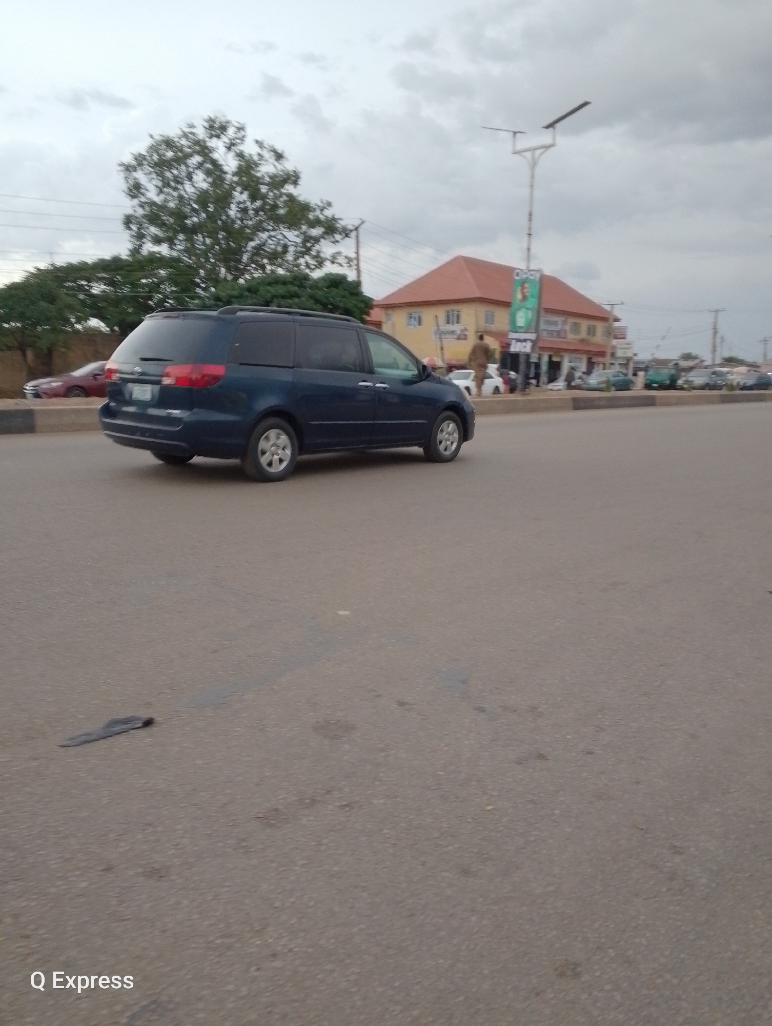 In an urban environment, likely Jos, Nigeria, under an overcast sky, a dark blue minivan drives on a paved road. The minivan, seen from a side angle and slightly from behind, is the most prominent subject in the foreground. A red car is visible on the left, further back. Buildings with orange roofs and some shops line the opposite side of the road. A street light pole with a solar panel is also present. The road is in a somewhat degraded condition with visible patches and debris. The overall atmosphere appears muted due to the cloudy weather.