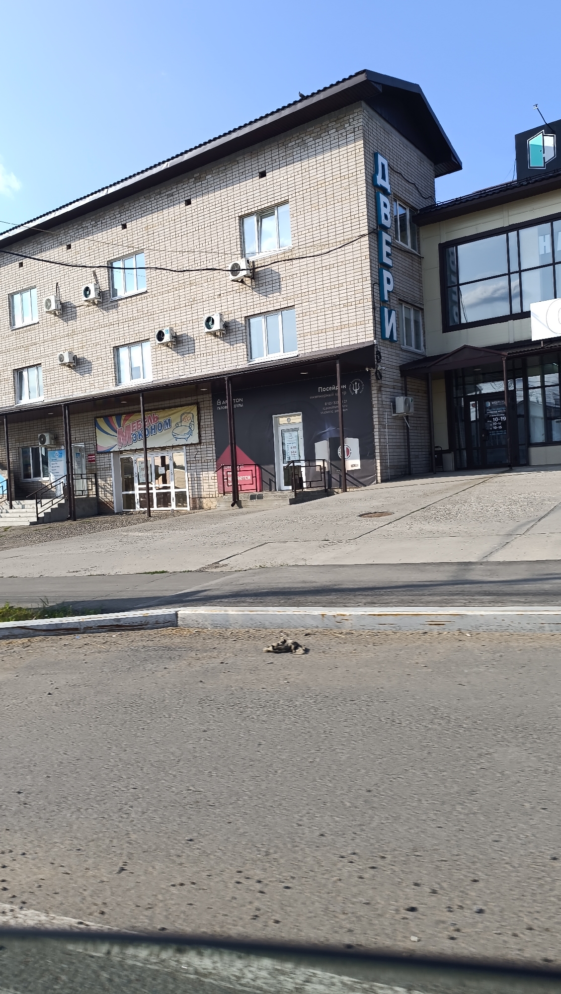 The image shows a multi-story brick building in Chaikovsky, Russia. The building has multiple windows and air conditioning units. On the side of the building, large blue letters spell out "ДВЕРИ" which translates to "DOORS" in Russian. Below the building, there are several storefronts, including one with a sign that reads "МЕБЕЛЬ ЭКОНОМ" (Furniture Economy) and another with a sign depicting an open door. The building is situated next to a road with a sidewalk and a patch of grass in the foreground. The sky is clear and blue.