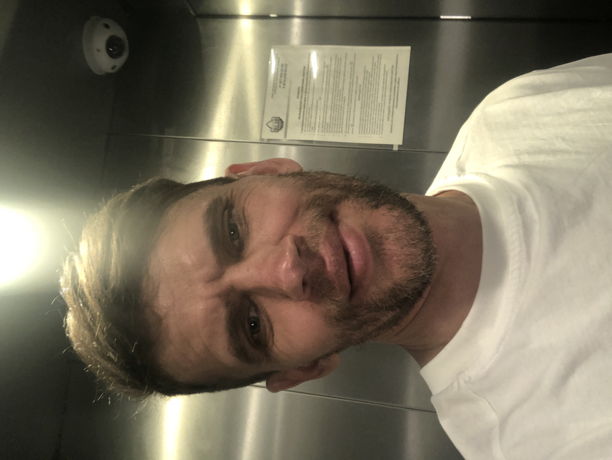 A man with short, light brown hair and stubble is taking a selfie in what appears to be an elevator. He is wearing a white t-shirt. In the background, the elevator walls are made of brushed metal, and there is a security camera mounted on the ceiling. A sign with Russian text is visible on the right wall.