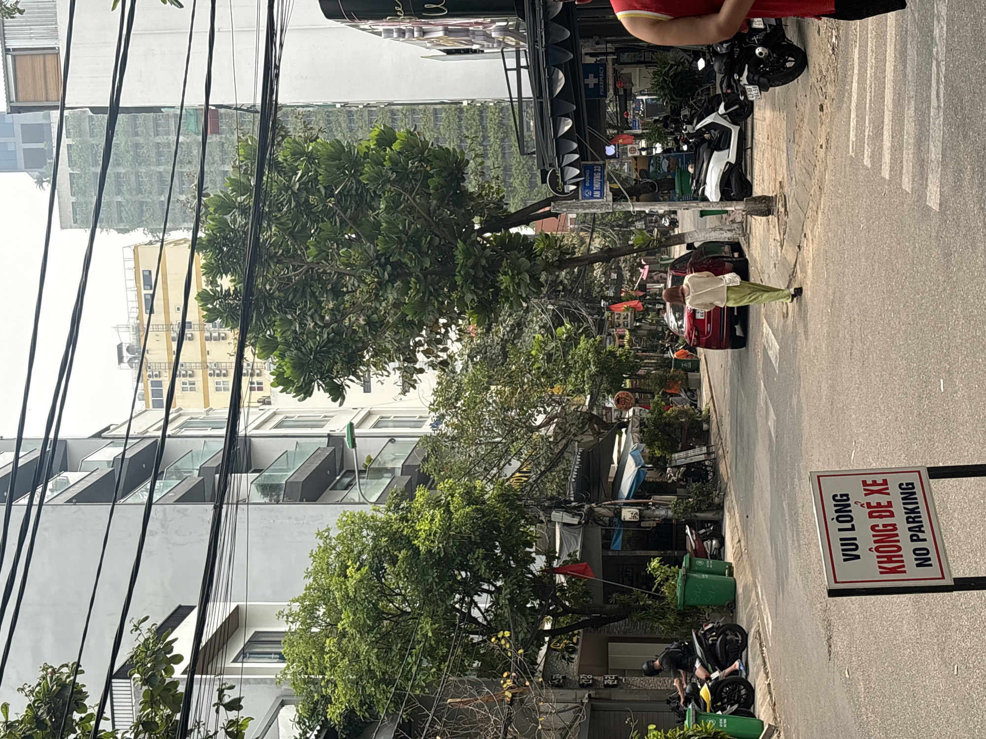 The image shows a street scene in Đà Nẵng, Vietnam. Tall buildings and power lines dominate the left side of the frame. Lush trees line the street. On the right, a woman is walking on the sidewalk next to a red car. A 'No Parking' sign stands on the edge of the sidewalk. Several parked motorbikes are adjacent to green trash bins. A pharmacy sign is visible in the background. The street sign 'ĐƯỜNG AN THƯỢNG 33' is visible above the pharmacy sign. A crosswalk is in the top right of the frame.