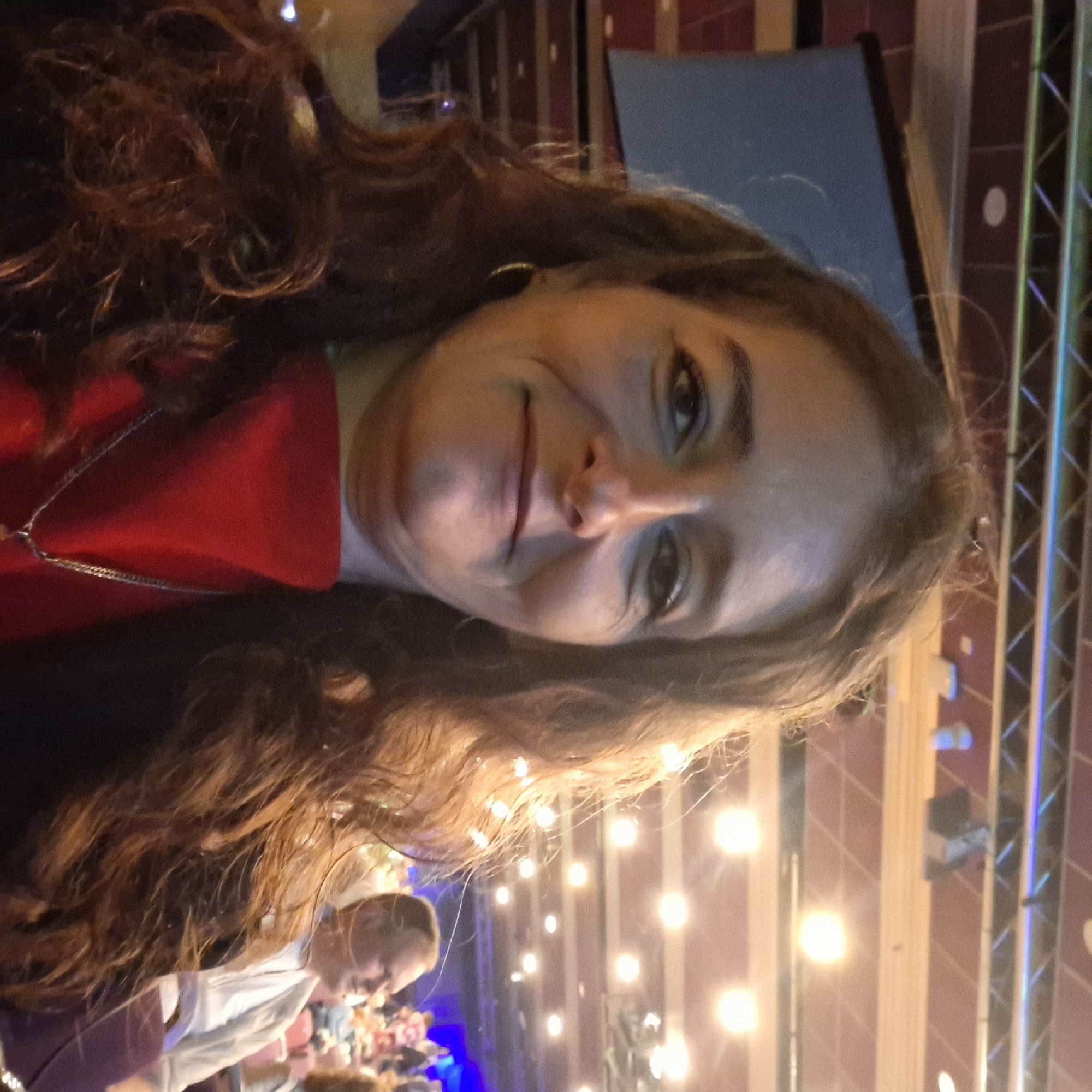 The image is a close-up, selfie-style photograph of a woman with long, wavy, reddish-brown hair, facing the camera. She has light skin, brown eyes, and is wearing a red top with a silver necklace. She is wearing a gold hoop earring in her visible left ear. In the background, there are rows of seats, some occupied by other people, suggesting an indoor event or performance space. Lights are visible on the ceiling. The overall impression is of a person attending an event. The image does not contain any recognizable landmarks or specific details that would indicate a location in Riga, Latvia.