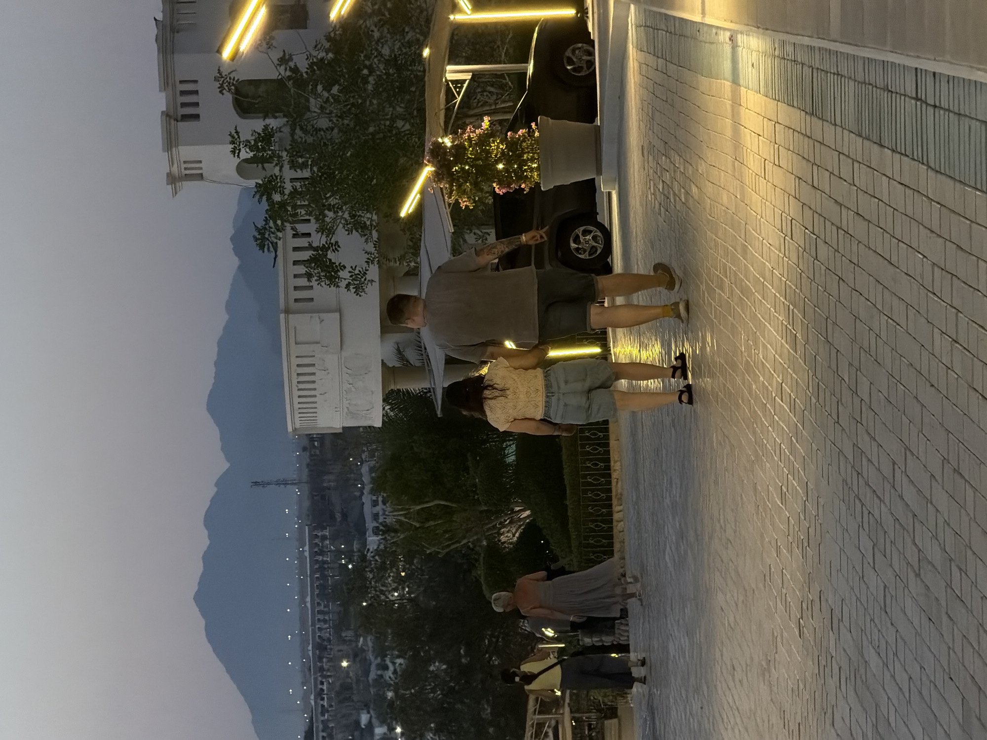 The image depicts an outdoor scene at dusk, possibly in Sharm El Sheikh, Egypt, given the mountainous backdrop and architecture. In the foreground, a paved walkway with subtle lighting leads the eye towards the distance. Several individuals are walking along this path. Closest to the viewer on the right, a person with short blonde hair and wearing a light yellow, lace-like top and denim shorts walks away from the camera. To their left, a couple walks hand-in-hand, also facing away. The man wears a grey t-shirt and dark shorts, while the woman is in a light-colored, textured top and light-colored denim shorts. Further down the path, two other individuals are visible, one in a grey dress and the other in a yellow top and dark pants, also walking away from the camera.
In the mid-ground, a dark-colored car is parked under a covered structure adorned with bright yellow lights. A potted plant with small pink flowers, also illuminated by lights, sits next to the car. Behind these elements, lush greenery and trees are visible, interspersed with the illuminated facade of a white, multi-story building with classical architectural features. In the background, silhouetted against the fading twilight sky, are undulating mountain ranges, suggesting a desert or arid environment. Lights from a distant city or resort are visible twinkling across the landscape. The overall atmosphere is serene and illuminated by artificial lighting, creating a warm and inviting ambiance.