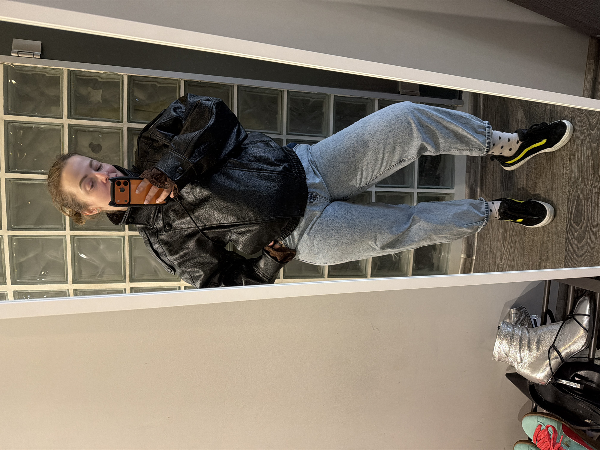 A female is pictured in a mirror selfie wearing a black leather jacket, light blue jeans, black shoes with yellow stripes, and polka dot socks. She is holding an orange smartphone. The background features a wall of glass blocks and a shoe rack with various shoes, including silver boots. There is no evidence that the image was taken in Moscow, Russia.