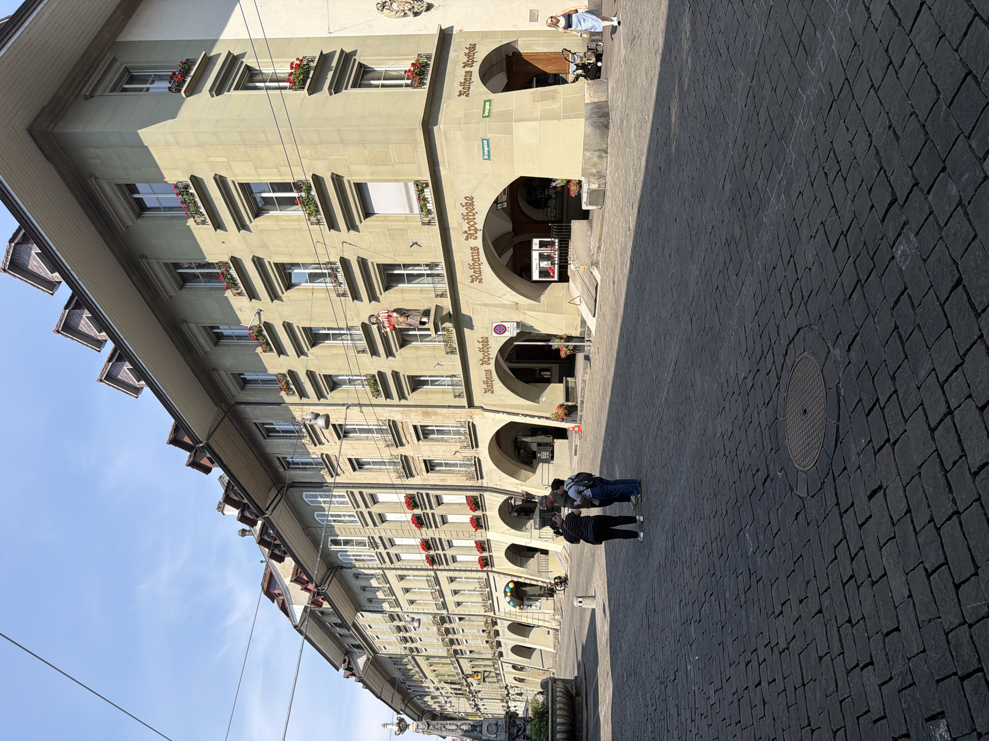 This image shows a street scene in Bern, Switzerland, specifically the Kramgasse in the Old Town. A long, pale yellow building with arched ground-level entrances lines the right side of the cobblestone street. Red flowers in window boxes adorn the upper floors of the building. In front of the building, three people are standing and talking. To the right, a woman is walking away from the camera. The street is paved with dark cobblestones, and shadows from the building stretch across the ground. The sky is clear blue with a few wispy clouds.