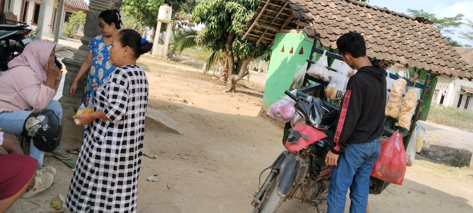 In Tanjung Ratu Ilir, Indonesia, three women and one young man are gathered around a motorbike outfitted with a display rack for goods. The motorbike is parked on a dirt road adjacent to a low-rise building with a tiled roof and green exterior. One woman, wearing a pink hijab and light-colored top, sits on a step, holding a phone to her ear. Another woman, in a blue dress with a cartoon print, stands nearby. A third woman, wearing a black and white checkered dress, holds two plastic bags containing yellow liquid and food items. The young man, wearing a black hoodie and blue jeans, stands by the motorbike, which has several plastic bags filled with food and snacks hanging from its rack, including packets of crackers. The environment appears to be a rural or semi-rural setting with some trees and vegetation visible.
