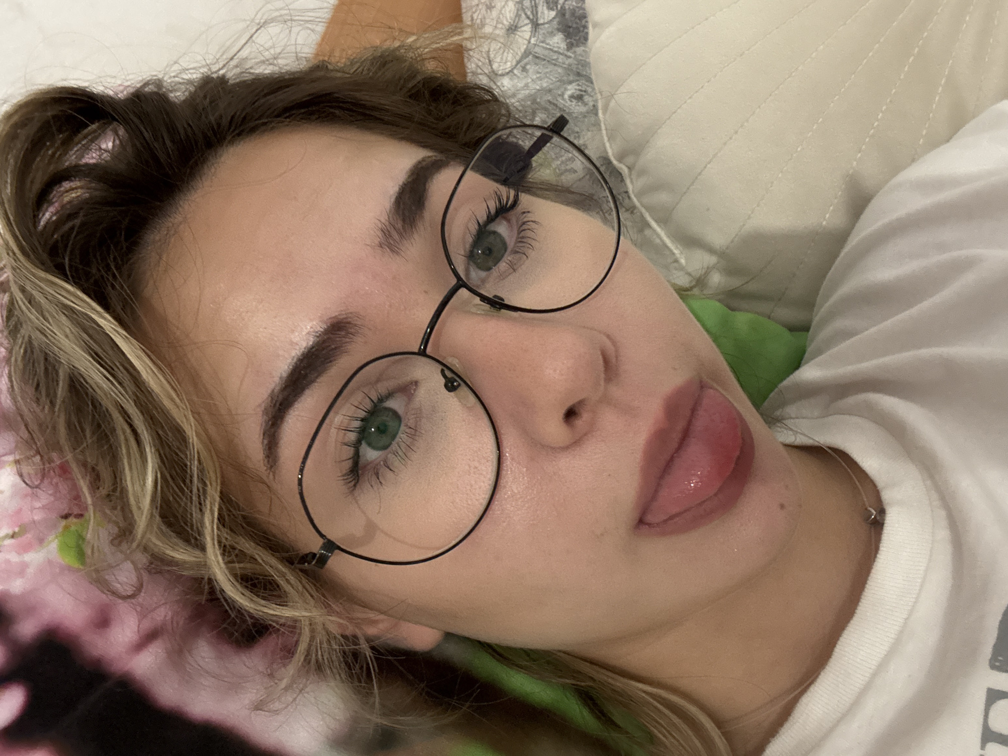 A close-up, eye-level selfie depicts a young woman lying down in an indoor setting, likely on a bed or sofa, in Blagoevgrad, Bulgaria. Her head is tilted, and she gazes towards the camera with a playful expression, her tongue extended slightly from her glossy, light pink lips.
She wears round, dark-framed eyeglasses that highlight her green eyes and long, well-defined eyelashes. Her eyebrows are dark and sculpted. Her hair features a mix of dark brown roots and blonde highlights, appearing in a relaxed, somewhat tousled style around her face and shoulders.
The woman is dressed in a white t-shirt, visible on the right side of the frame, and a thin necklace with a small, metallic charm. The background consists of soft furnishings, including a light beige or off-white quilted pillow behind her head and a glimpse of a green fabric object beneath it. A patterned fabric with dark and pink floral designs is visible on the left, contributing to a cozy and personal atmosphere. The lighting is soft and even, suggesting an indoor environment without harsh direct light. No visible text is present in the image.