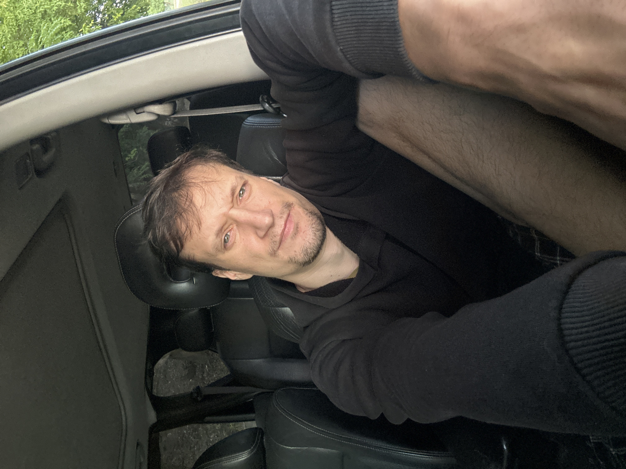A man is seated in the driver's seat of a car, with the camera angled to capture a selfie. He has brown hair, fair skin, and a light beard. He is wearing a black hoodie. The interior of the car, with its black leather seats and sunroof, is visible. Outside the car, green trees can be seen. The scene appears to be outdoors, likely in a rural or suburban setting, and there is no indication of Makiv, Ukraine.