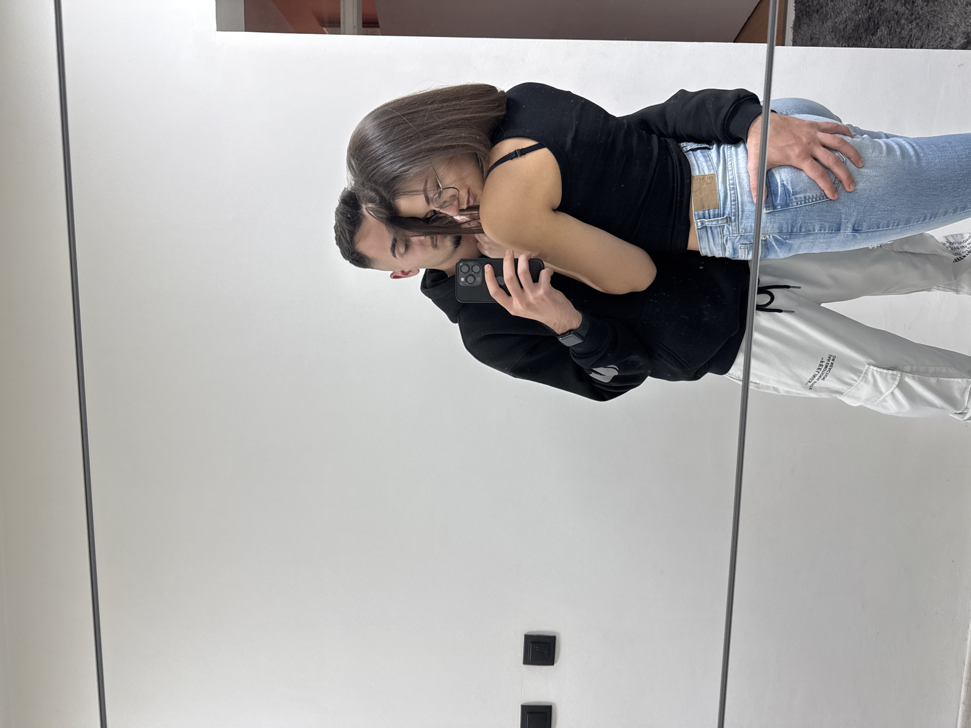 A couple is captured in a mirror selfie. The man is wearing a black hoodie and white pants and holding an iPhone. The woman is wearing a black top and light blue jeans, leaning into the man. The background appears to be a simple white wall with electrical outlets. The location is implied to be in the city of Sofia, Bulgaria.
