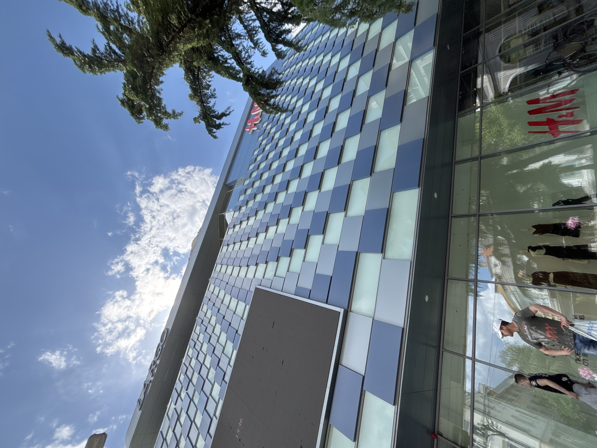 The image captures the modern facade of the LARGO shopping center in Blagoevgrad, Bulgaria. The building features a distinctive exterior composed of alternating light and dark blue rectangular panels, arranged in a checkered pattern. A large digital screen is positioned on the facade. The red H&M logo is visible on the building. Reflections in the glass windows show people and the surrounding environment. A tree with dense foliage overhangs the top of the building. The sky is visible, with scattered clouds and blue sky. Two people are visible through the reflections of the building.