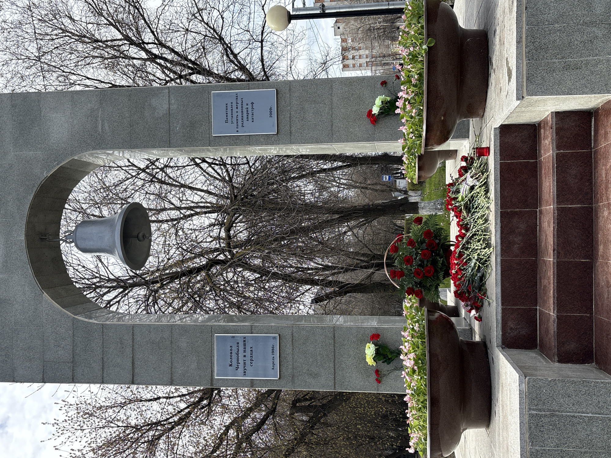 The image shows a memorial monument in Ulyanovsk, Russia, commemorating victims of radiation accidents and disasters, including Chernobyl. The monument consists of a stone arch with a bell suspended in the center. Two plaques are attached to the arch. The upper one, translated, reads, "A monument erected in memory of the victims of radiation accidents and catastrophes," followed by "2009." The lower plaque reads, "The bell of Chernobyl sounds in our hearts," and "April 1986." Two decorative brown urns are partially visible, flanking the memorial. Red and white flowers are placed near the base of the monument and in the urns. A set of steps leads up to the memorial. Bare trees are visible in the background, suggesting the season is either late autumn or early spring. In the distance, buildings can be seen. A lamppost stands nearby.